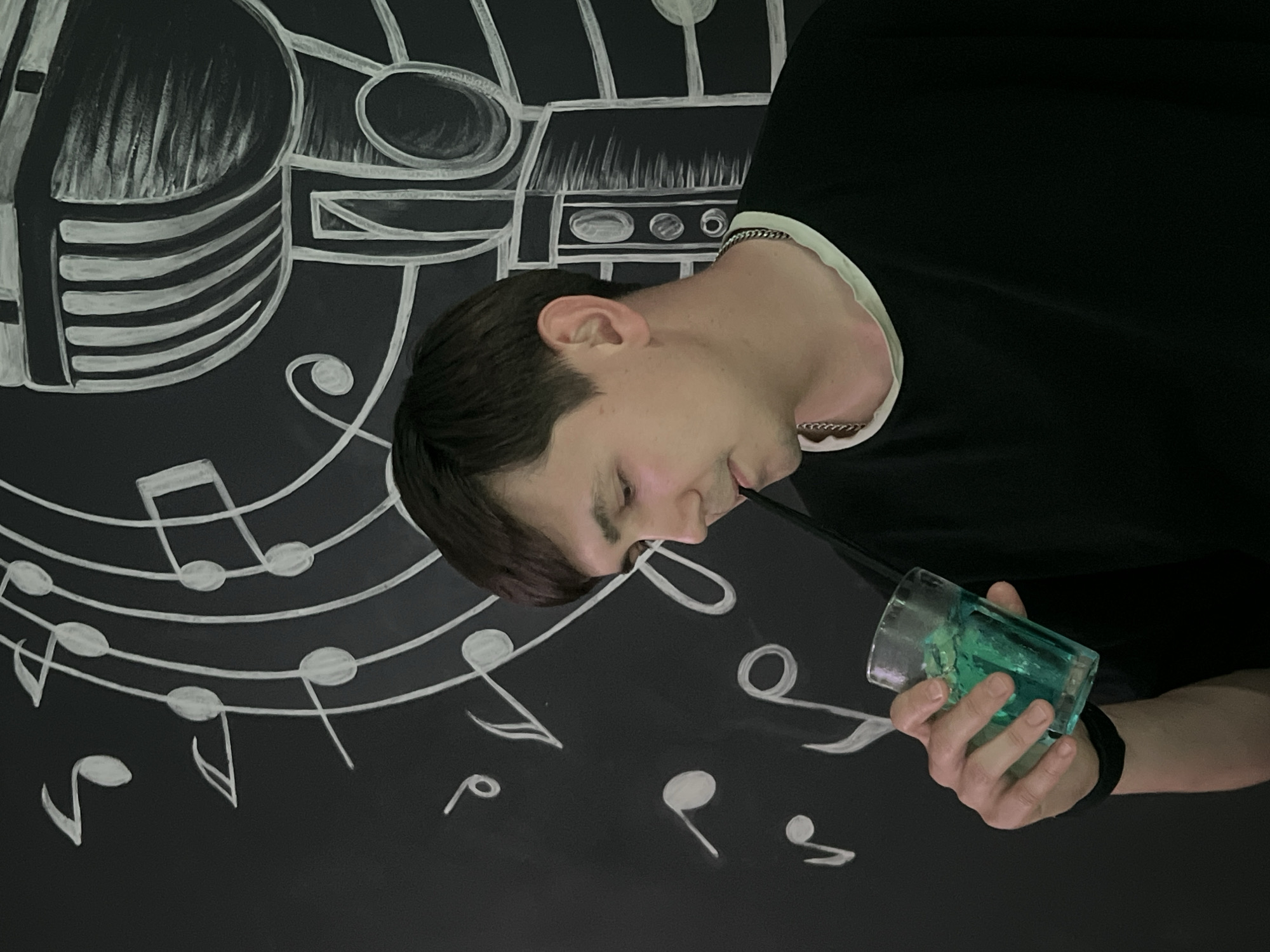 A young man with short dark hair is holding a glass of a blue-green beverage with a straw in his right hand, to his lips. He is wearing a black t-shirt with a light-colored trim around the neck and a silver chain necklace. The man is positioned in front of a dark wall with a chalk drawing of musical notes and a microphone. The background suggests an indoor setting, possibly a bar or music venue. The image does not provide any information about the city of Chaikovsky, Russia.