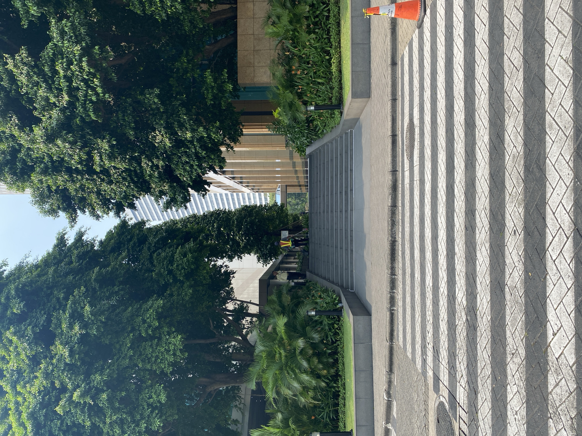 The image depicts a pedestrian crossing in front of a modern building with extensive landscaping and a grand staircase. The crossing is paved with interlocking bricks and marked with white stripes. To the right of the crossing, an orange traffic cone is placed. The staircase is wide and made of grey stone, leading up to the building's entrance. Flanking the staircase are lush green plants, including palm trees and flowering plants. Two individuals, possibly security personnel due to their attire (one in a light-colored uniform, the other in a bright vest), are visible at the top of the stairs. Behind the landscaping and building, a tall, modern skyscraper rises into the clear sky. The scene is set in a city environment, with the architecture and landscaping suggesting a well-maintained urban space. The location is identified as Central Jakarta, Indonesia.