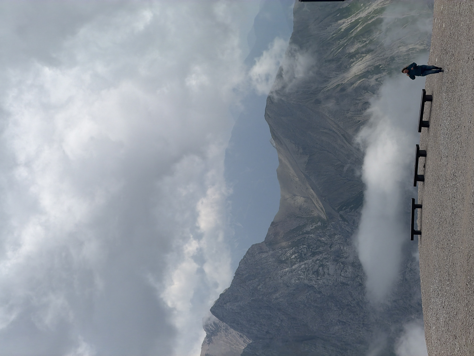 The image depicts a dramatic mountainous landscape with a person seated on a bench. The scene is set in a high-altitude, mountainous region, likely Garmisch-Partenkirchen, Germany, given the context. The weather appears to be overcast with thick clouds obscuring parts of the sky and rolling in around the peaks, creating a sense of moody atmosphere.
The primary subject is a person, dressed in dark clothing, sitting on a dark-colored bench. The person is facing away from the camera, looking out at the vast mountain scenery. Their posture suggests a moment of contemplation or rest. In the foreground, there are two dark benches positioned along a textured, gravelly surface that appears to be a walkway or viewing platform.
The background is dominated by imposing, rugged mountain ranges. The peaks are sharply defined, with steep inclines and rocky textures visible. Clouds are interspersed among the mountains, sometimes shrouding entire sections and adding a sense of depth and mystery to the scene. The lighting is diffused due to the cloud cover, indicating it is likely daytime, though not a bright, sunny day. The overall mood is one of awe and perhaps a touch of solitude, enhanced by the scale of the mountains and the solitary figure. There is no visible text in the image.