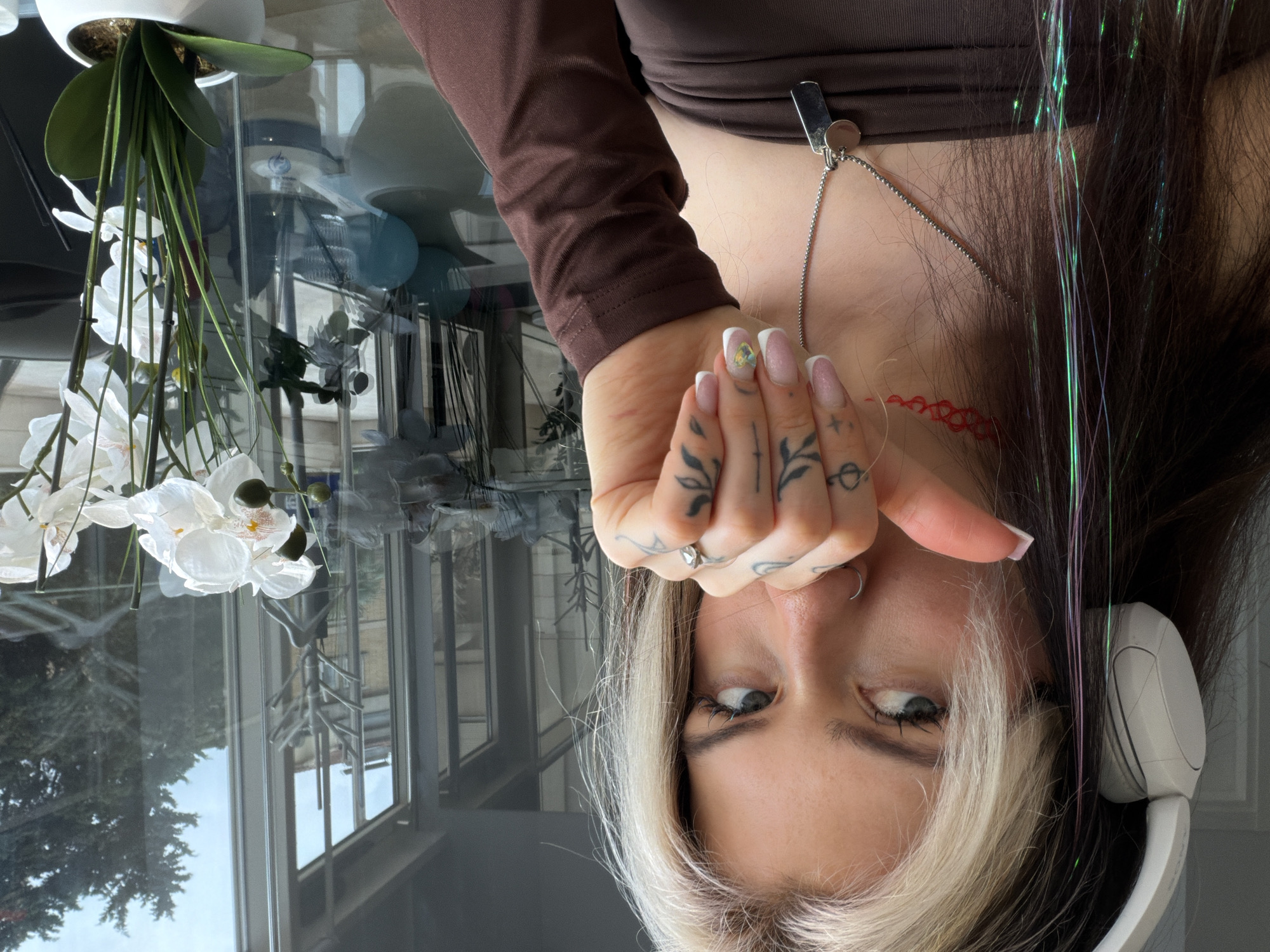 The image captures a close-up of a person looking directly at the camera, with their head slightly tilted upwards. Their gaze is intense and direct. They have dark hair with prominent blonde streaks and wear white over-ear headphones on their left ear. Shimmering green and purple strands, likely hair tinsel, are visible within their dark hair. The person is wearing a brown long-sleeved top and a silver clip-on necklace.
Their right hand is raised near their face, adorned with small dark tattoos on the fingers and knuckles, and a ring. The fingernails feature a French tip design, with one nail displaying an iridescent gem. A distinctive red, braided pattern tattoo is visible on their neck.
The background shows a large window revealing an outdoor scene with trees and parts of a building under clear daytime skies. An artificial white orchid plant in a white pot rests on a reflective glass surface, possibly a table, which displays subtle reflections of the room and outdoor environment. No text is visible in the image. The scene is set indoors in Budva, Montenegro.