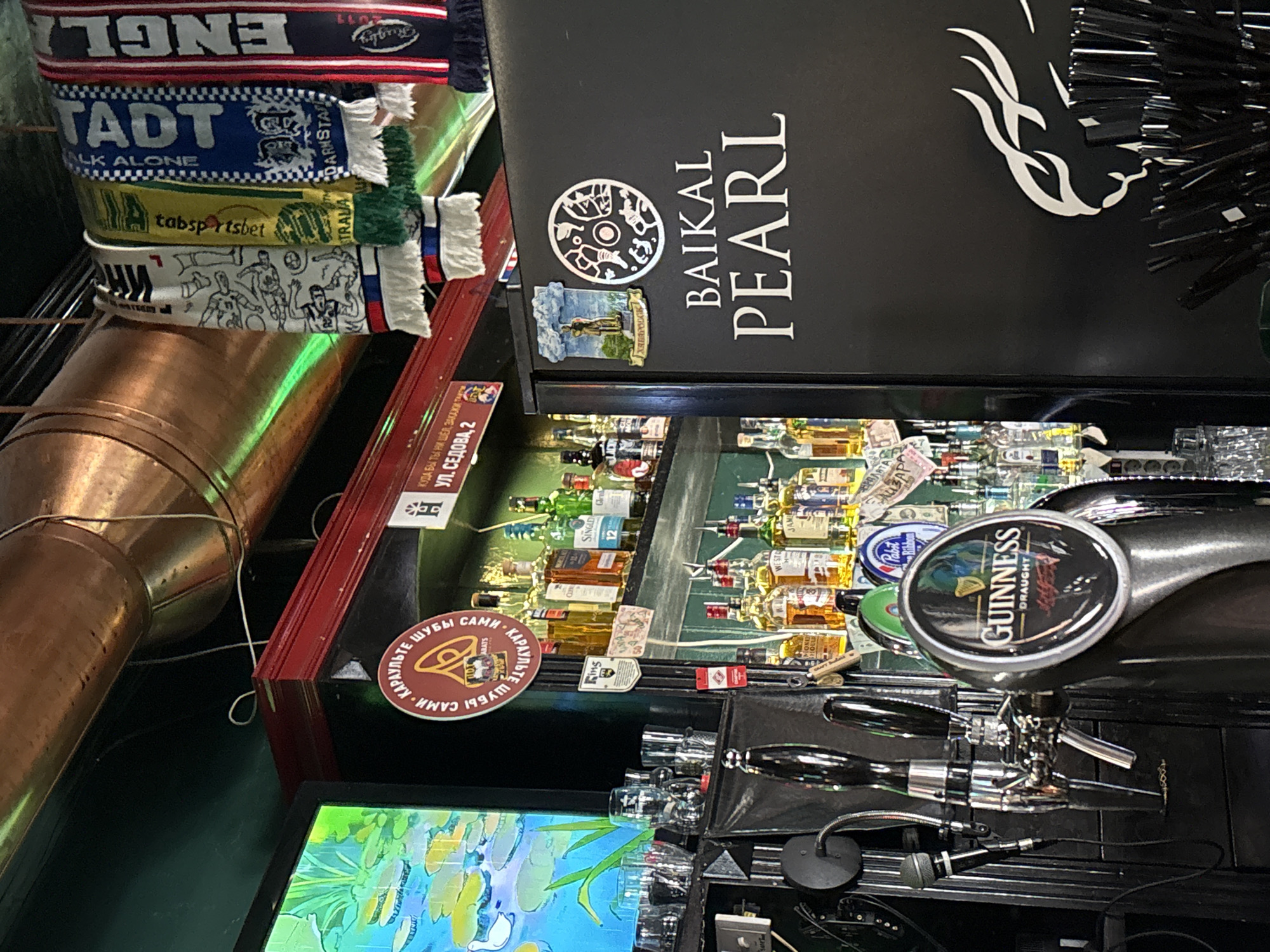 The image depicts the interior of a bar. A Guinness tap is visible, along with an assortment of alcohol bottles displayed on a shelf, some adorned with money. Scarves representing different soccer teams are stacked on the left. The signage "BAIKAL PEARL" is also visible. A sign reads "УЛ. СЕДОВА, 2" indicating the location is Sedova Street, number 2. A sign mentions "КАРАУЛЬТЕ ШУБЫ САМИ - КАРАУЛЬТЕ." The bar is likely in Irkutsk, Russia.