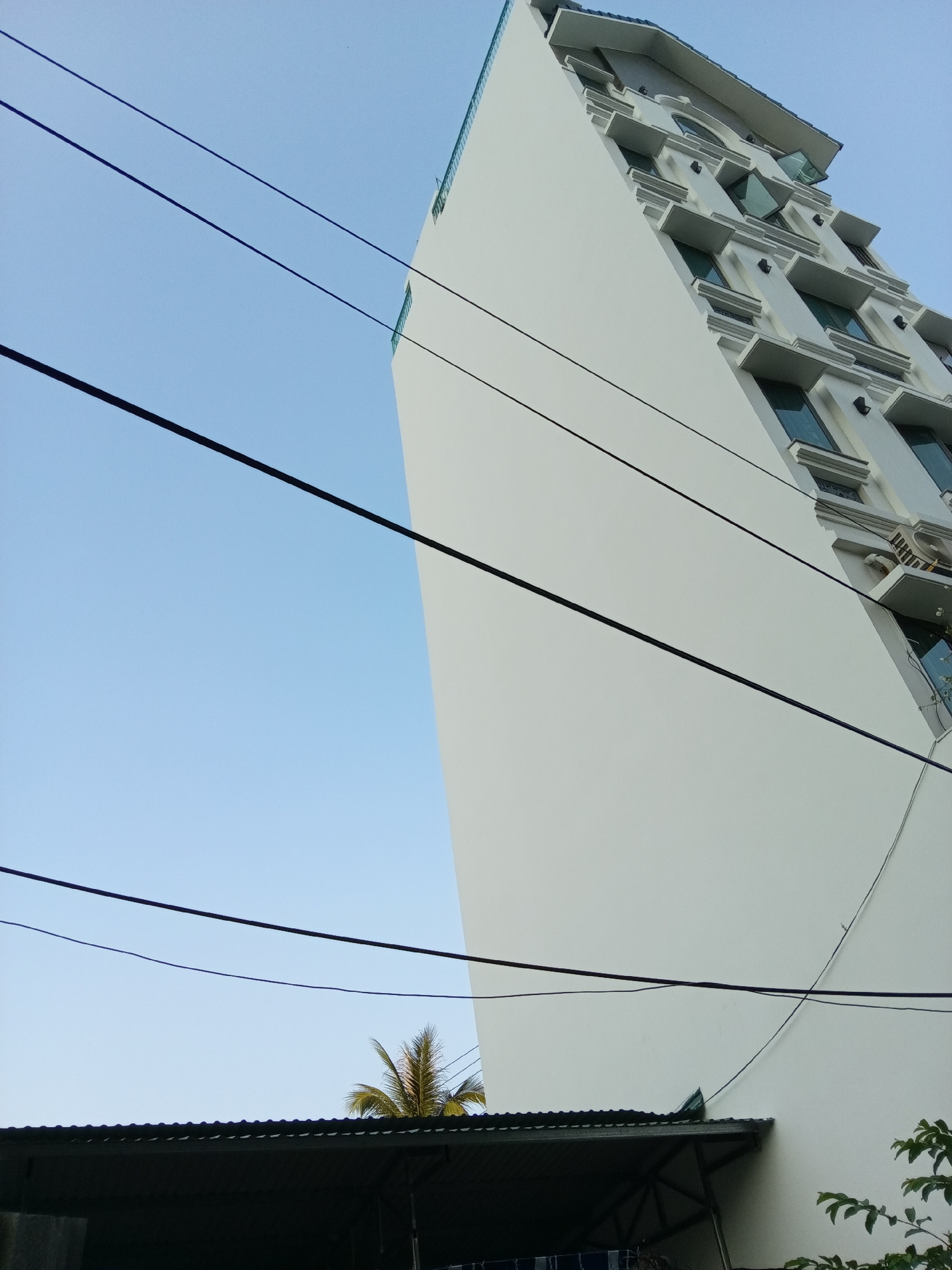 A low-angle shot reveals a tall, white building under a clear blue sky in Bắc Nha Trang, Vietnam. The building, extending upwards and slightly to the right, features rows of rectangular windows with decorative ledges. Power lines crisscross the sky in the foreground. Below, the corner of a corrugated metal roof is visible, with a partial view of a palm tree and some foliage to the side. Laundry hangs underneath the shelter.