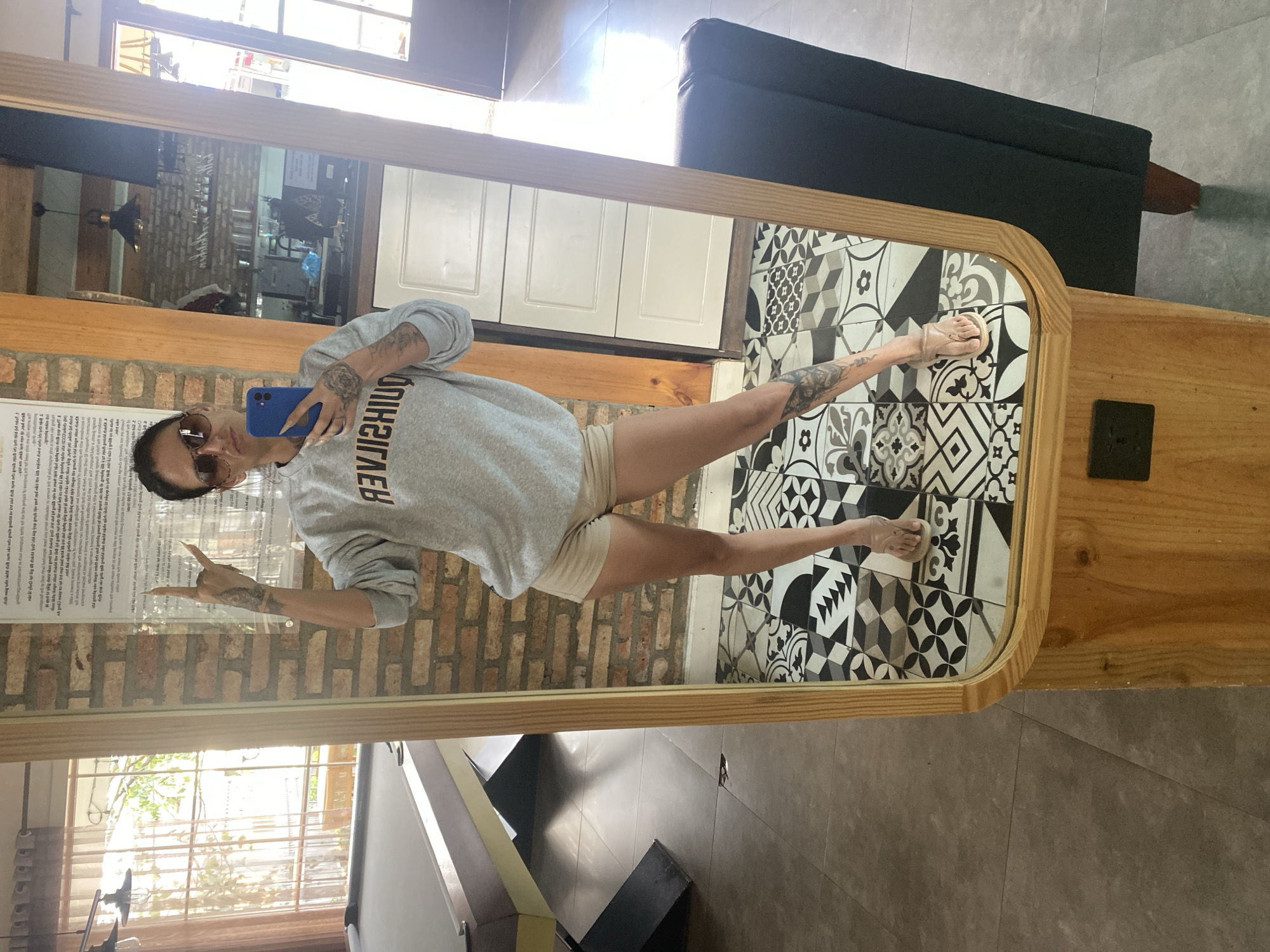 A woman is taking a mirror selfie in a room. She is wearing a gray sweatshirt, beige shorts, and sunglasses. She is holding a blue phone in her left hand and making a peace sign with her right hand. The room has a pool table in the background and a black armchair. The floor is tiled with a black and white geometric pattern. The image is taken from a low angle. The location is not specified in Da Lat, Vietnam.