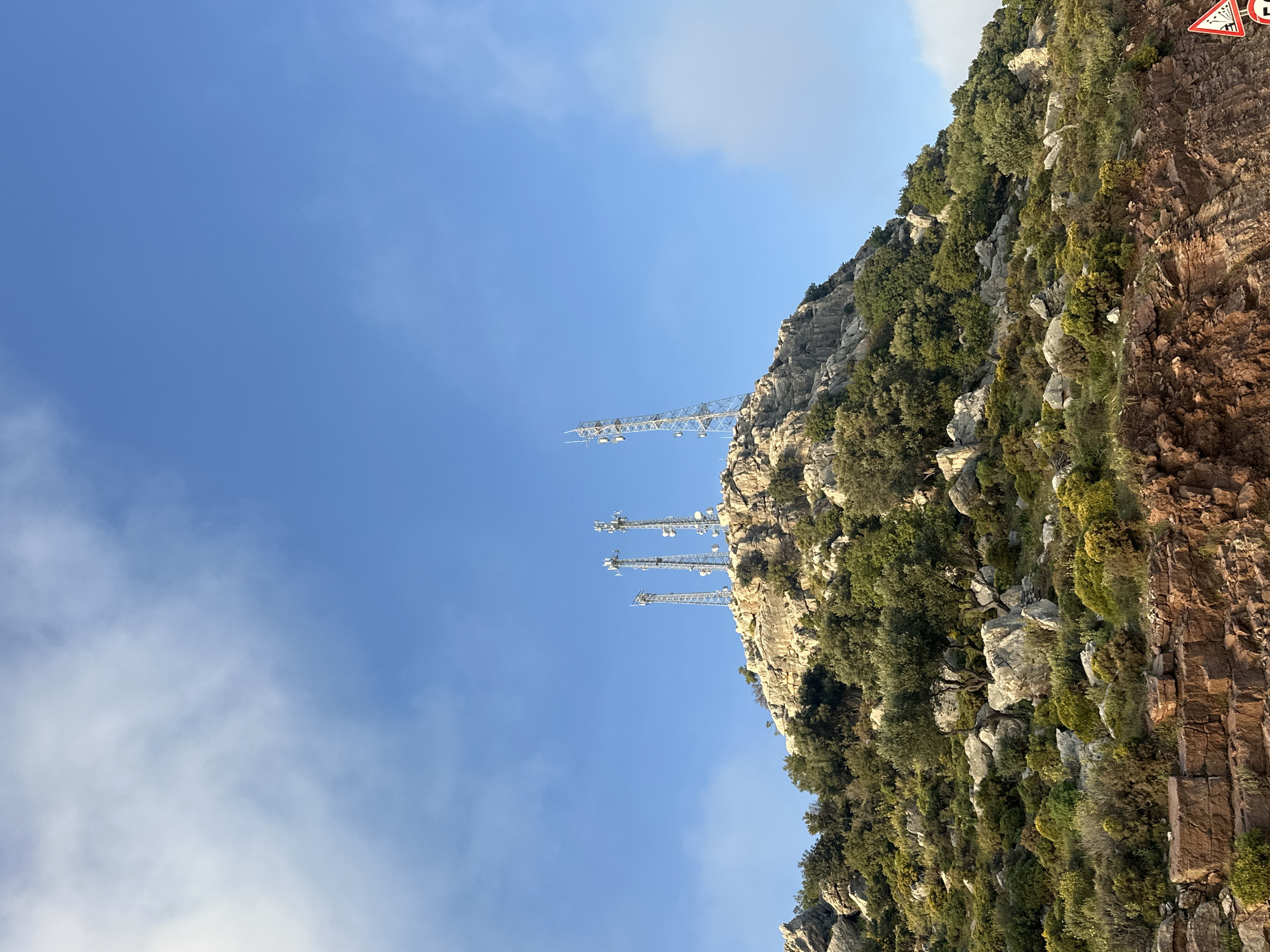 This media file presents a rugged, rocky mountain face or cliff under a bright, clear blue sky dotted with scattered white clouds. The landscape is characterized by prominent light-colored rock formations interspersed with patches of green vegetation, including bushes and small trees, suggesting a dry or Mediterranean climate, consistent with the Fethiye region of Turkey.
Perched on the crest and upper slopes of the mountain are several tall, grey metal lattice structures, identified as communication towers or telecommunication masts. At least four distinct towers are visible, with one appearing as a substantial horizontal lattice framework extending outwards from the cliff face.
In the upper right corner, a red and white triangular road sign is partially visible. It displays the universal black symbol for falling rocks, indicating a potential hazard or the proximity of a road below the cliff. On the upper right section of this sign, the letter "C" and another partial letter, possibly "L," are visible in white against a dark background.
The time of day appears to be daytime, with ample sunlight illuminating the scene and casting soft shadows, suggesting either morning or afternoon. There is no explicit activity or event taking place; the image is a static landscape shot highlighting the interplay between nature and modern infrastructure.