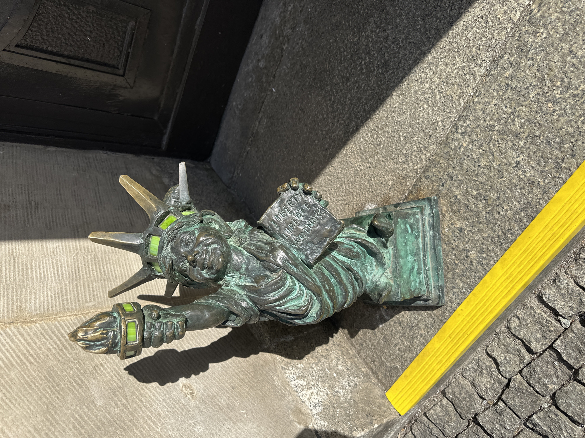 A metal statue of a dwarf dressed as the Statue of Liberty sits at the edge of a stone staircase near a black door. The dwarf wears a crown with green inserts, holds a tablet in his left hand, and raises a torch in his right hand. A yellow safety strip runs parallel to the dwarf on the edge of the staircase. The statue is likely located in Wrocław, Poland, where numerous dwarf statues are found throughout the city.