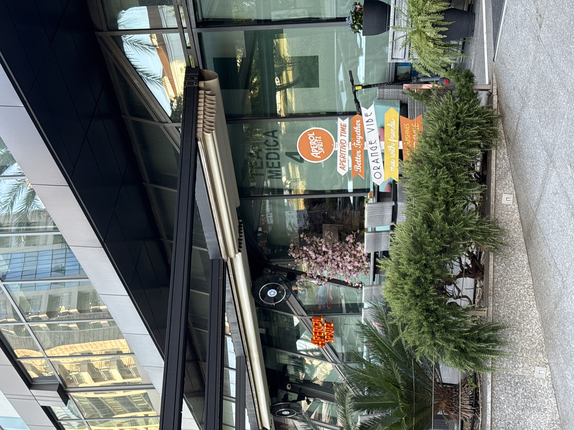 This is an exterior view of a building, likely a café or restaurant, in Budva, Montenegro. The building has a modern architectural style with glass windows reflecting the surrounding environment, including palm trees and other buildings. A retractable awning shades the entrance.
Promotional signs in front of the building advertise "TEA MEDICA," "APEROL SPRITZ," "APERITIVO TIME," "ORANGE VIBE," and a message about mixing with friends. A small scooter is parked near the entrance. Decorative plants and trees line the sidewalk, adding greenery to the urban landscape. The foreground shows a portion of the paved sidewalk.