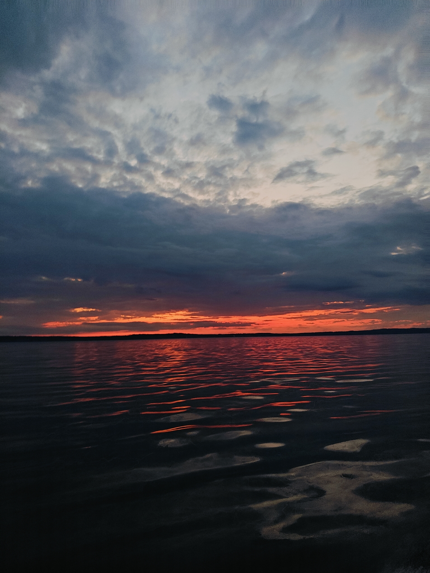This is a landscape photograph taken at sunset over a body of water, likely the Kama River reservoir, near the city of Chaykovsky, Russia. The sky is filled with dramatic clouds, transitioning from dark, moody grays at the top to vibrant oranges and reds along the horizon where the sun is setting. The water in the foreground reflects these fiery colors, creating streaks of light on its dark, rippling surface. The distant shoreline is a dark silhouette of trees, indicating a natural or semi-natural environment. There are no people or man-made structures clearly visible in the image, emphasizing the natural beauty of the scene.