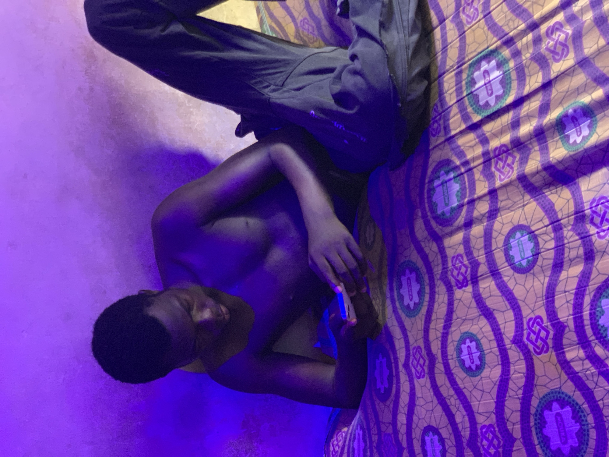 A shirtless man, likely in Lokoja, Nigeria due to the presence of patterned cloth typical of that region, is seated on a bed, wearing dark-colored pants. The lighting is primarily purple, casting strong shadows. The man appears to be looking at a rectangular object held in his hands, possibly a phone. He is situated near a wall painted a light color. The bed has a patterned yellow and purple fabric covering it. The overall setting is simple and domestic.