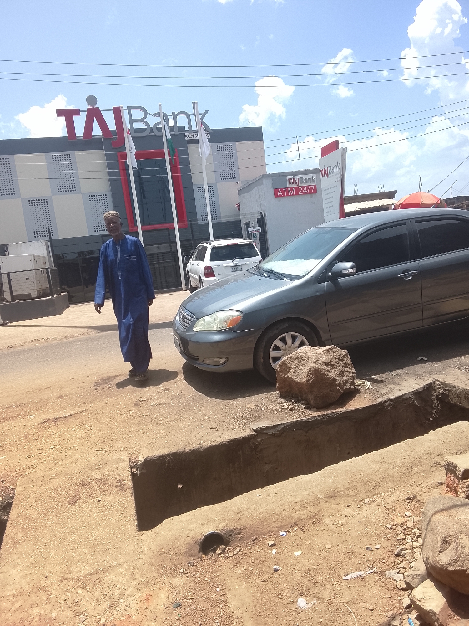 The image shows a street scene in Jos, Nigeria, in front of a Taj Bank branch. A man in a long blue robe is walking on the left side of the street. A gray Toyota Corolla is parked on the right side of the street, next to a large rock. A white Toyota Highlander is parked behind the Corolla. The street is unpaved and has a deep trench. A Taj Bank ATM booth is visible in the background to the right of the bank building. The sky is partly cloudy.