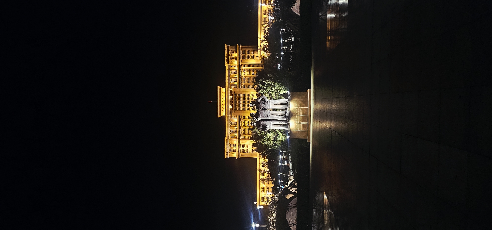 In the city of Beibei, China, at night, a wide shot captures a grand, illuminated building in the background. The building is a multi-story structure with a symmetrical facade, featuring numerous windows and decorative elements, all bathed in a warm, golden light emanating from external fixtures. In the foreground, centered on a raised pedestal, stands a bronze statue of three men. The men are depicted in uniform, with one figure pointing forward. They are positioned against a backdrop of manicured trees and shrubs, also illuminated, creating a dramatic contrast with the dark night sky. The ground in front of the statue is paved with large, square tiles, reflecting the light from the building and the statue, suggesting a damp or wet surface. The overall scene evokes a sense of solemnity and grandeur, likely a public monument or landmark.
