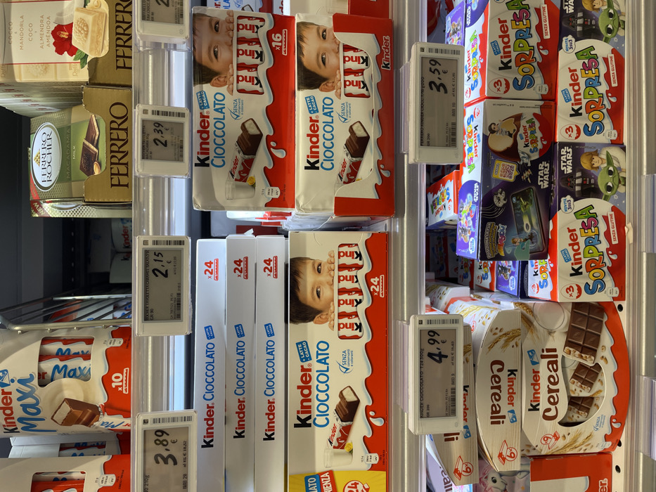 This image depicts a brightly lit confectionery aisle within a retail store in Monsummano Terme, Italy. The scene focuses on multiple shelves fully stocked with various Kinder brand chocolate products, alongside other Ferrero items.
The top shelf on the left displays Kinder Maxi chocolate bars in white and red packaging, indicating "x10" units, priced at 3.89€. Adjacent to this, in the upper right quadrant, are Ferrero Rocher Macadamia Milk chocolates in a gold-brown box, priced at 2.39€, and a white-packaged Ferrero product labeled "Coco & Mandorla."
The middle shelves are predominantly filled with stacks of Kinder Cioccolato boxes. These white boxes feature a child's face peeking over a row of chocolate bars, with "LATTE + CACAO" and "SENZA GLUTINE" labels. Some boxes indicate "x24" units, with one shelf displaying a price of 4.99€ for a Kinder Cioccolato "T24 500G." Other stacks of Kinder Cioccolato are marked "x16" units.
The bottom shelf presents Kinder Cereali chocolate bars in white and brown packaging, showing chocolate bars with cereal grains. To the right, multiple boxes of Kinder Sorpresa (surprise eggs) are visible. These boxes are predominantly white, red, and blue, labeled "SENZA GLUTINE," and some feature "Star Wars" branding with characters like Yoda. A digital price tag above these boxes displays 3.59€ and identifies the product as "FERRERO KINDER CEREAL T/10/200," despite its placement above Kinder Sorpresa.
All product prices are displayed in Euros on electronic white digital tags. The shelves are metallic and well-organized. No individuals are visible in the scene.