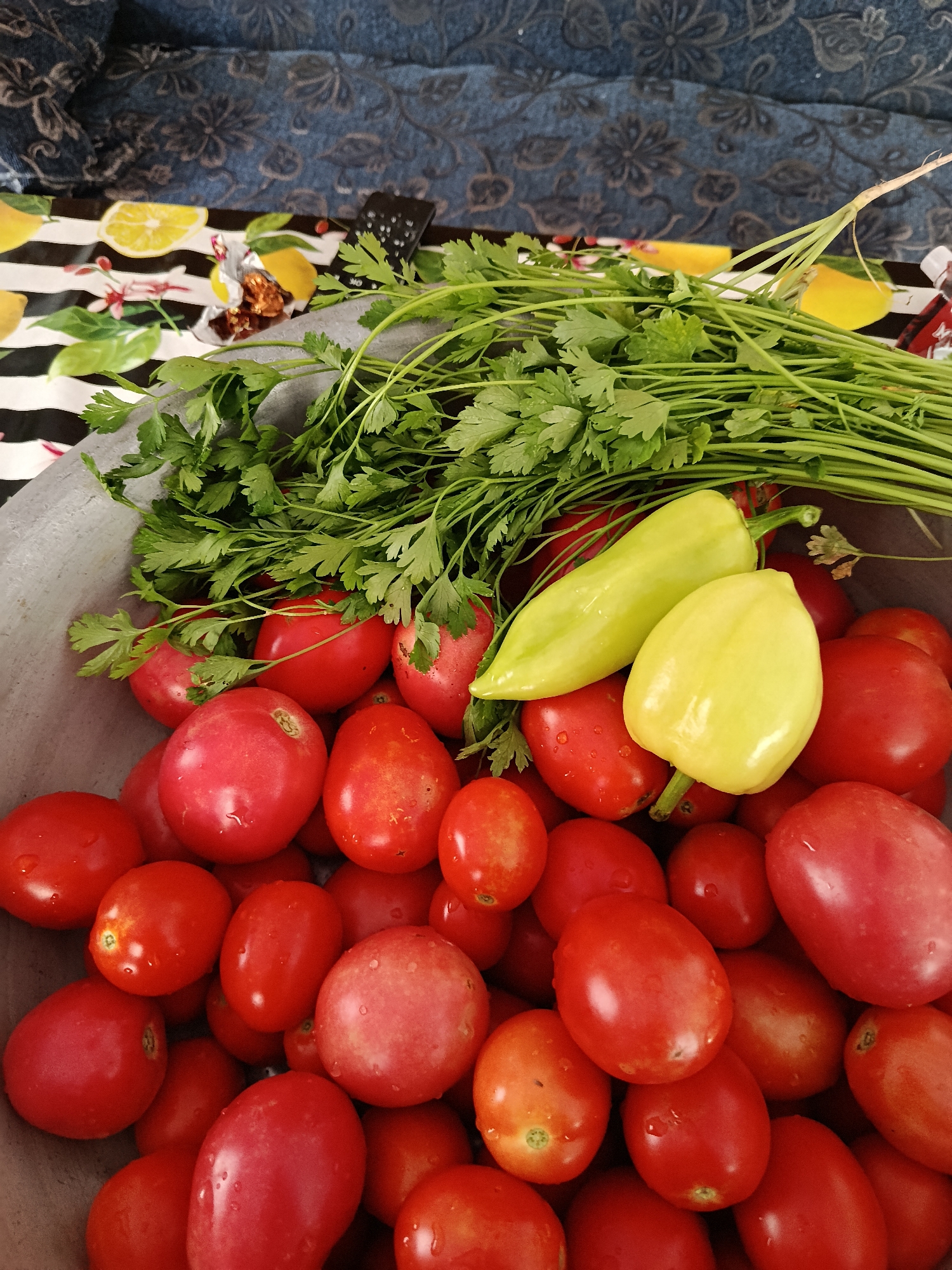 A close-up, overhead view of a large metal bowl filled with ripe red tomatoes and two pale green bell peppers. A bunch of fresh parsley is draped over the vegetables. In the background, a patterned fabric and a table runner with a lemon motif are visible. The scene appears to be set indoors, possibly in a kitchen or dining area. The city of Койлык, Kazakhstan, is mentioned in the prompt, but there is no visual information in the image to support or refute its location.