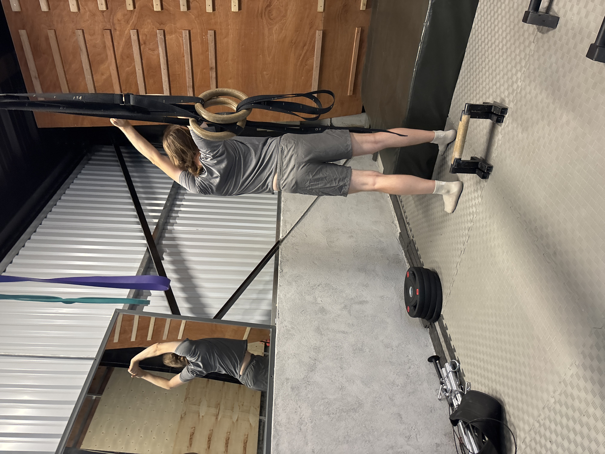 In a gym setting, a person is performing an exercise using gymnastic rings. The individual is wearing a grey t-shirt and grey shorts with white socks. Their body is positioned horizontally, with their hands gripping the rings and their feet extended behind them. The scene is indoors, with wooden panels and metal structures visible in the background. There are also exercise equipment like weights and push-up bars present. The overall lighting suggests it could be daytime. The environment appears to be a functional training space, possibly a CrossFit gym or a similar facility.