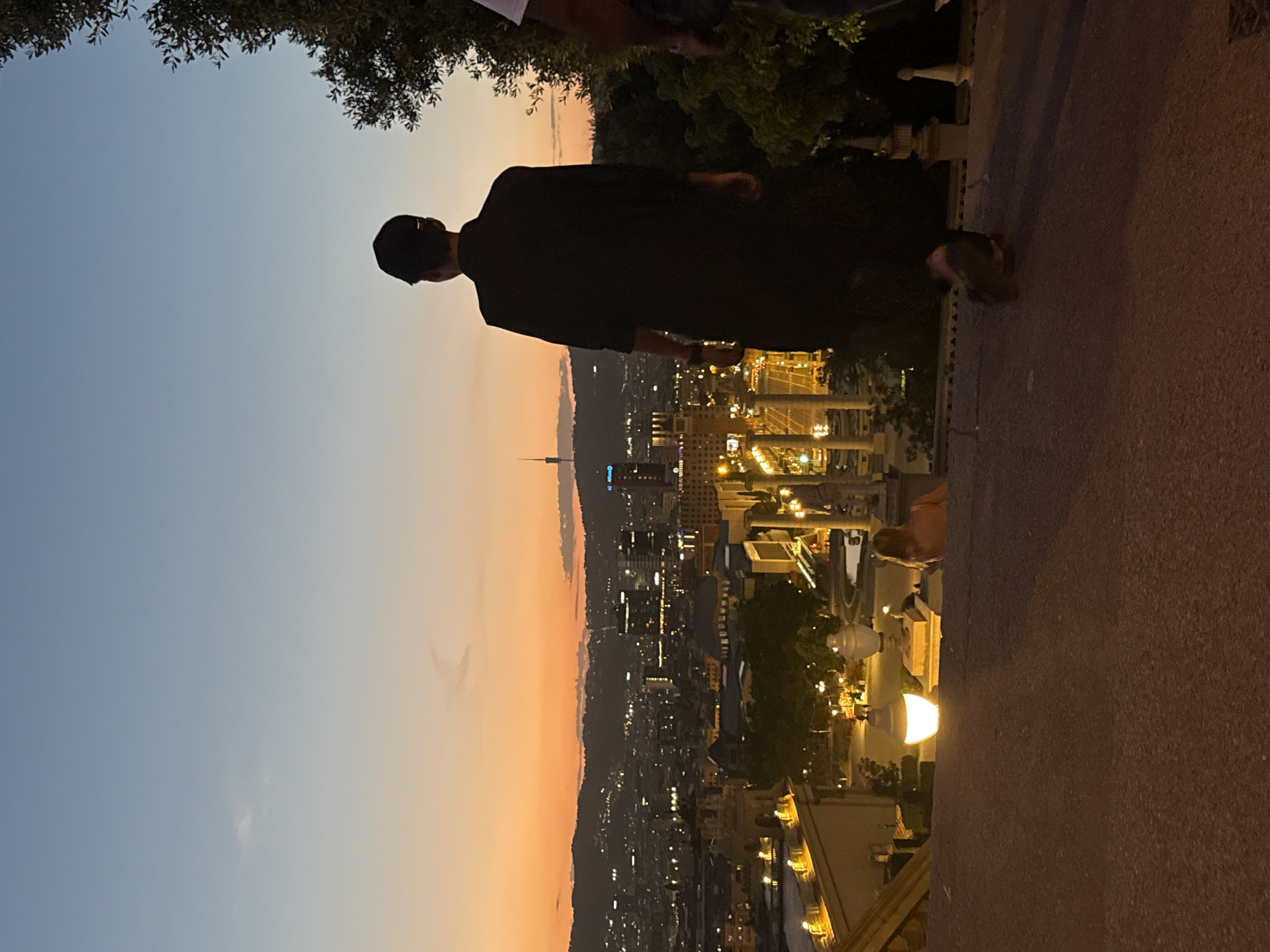 The image depicts a sunset view of Barcelona, Spain, taken from a vantage point overlooking the city. In the foreground, a person wearing a dark cap and black clothing stands with their back to the camera, facing the cityscape. To the right of this individual, a portion of another person is visible, also facing away from the camera.
The city below is illuminated by a multitude of lights, suggesting it is dusk or early evening. A prominent tower with a slender spire rises from the city on the left side of the frame. The sky transitions from a soft orange hue near the horizon to a pale blue overhead, with subtle clouds.
In the mid-ground, there are architectural elements of what appears to be a public space, including columns and ornate lamps, one of which is lit. Further into the city, buildings of various heights and styles are visible. A person in an orange top is seated in the middle ground, also facing away from the camera. The scene is framed by trees on the right side. The foreground consists of a textured surface, likely a path or stairway.