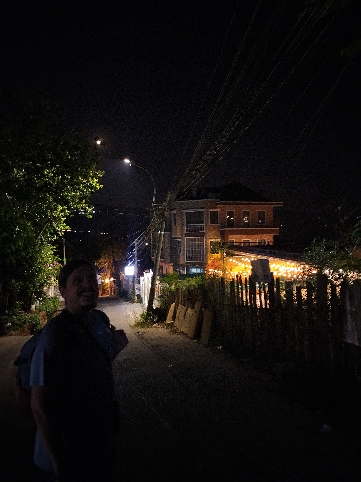 The image captures a dimly lit street scene at night in Da Lat, Vietnam. A man with a backpack stands on the left side of the frame, his face visible in the limited light. Overhead power lines crisscross the sky.
The street stretches into the distance, illuminated by a street lamp and lights from buildings. On the right, a wooden fence borders a property with string lights, suggesting a restaurant or similar establishment. A multi-story building is visible beyond the fence. The road is paved, and vegetation grows on either side. The sky is dark, with a hint of moonlight.