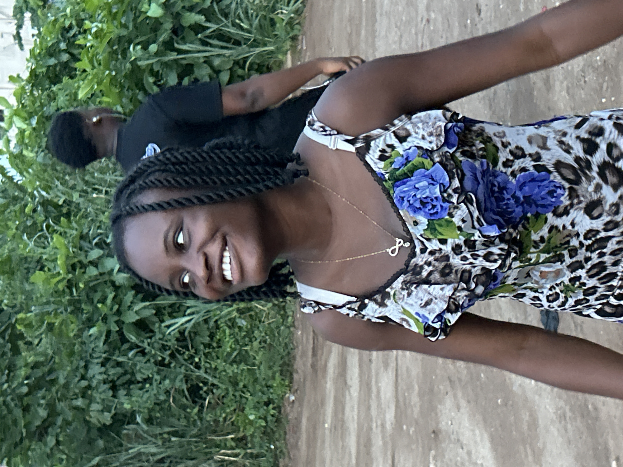 This outdoor image features two young Black individuals. The primary subject, a young woman, is positioned prominently in the foreground, turning to face the camera with a wide, joyful smile that reveals her white teeth. Her hair is styled in dark braids. She wears a sleeveless top featuring a distinctive leopard print combined with vibrant blue floral patterns. Around her neck is a delicate gold chain necklace with an infinity symbol pendant.
Behind her, partially obscured by green foliage and the primary subject, is another person, seen from the back, wearing a plain black t-shirt. This individual appears to be walking or standing casually.
The setting is an outdoor environment characterized by lush green foliage, likely bushes or tall grass, on the left side and in the background. To the right, a dirt or unpaved path or road is visible. The lighting suggests it is daytime. The overall atmosphere conveyed by the main subject's expression is one of happiness and approachability. The scene takes place in Ihiagwa, Nigeria. No visible text is present in the image.