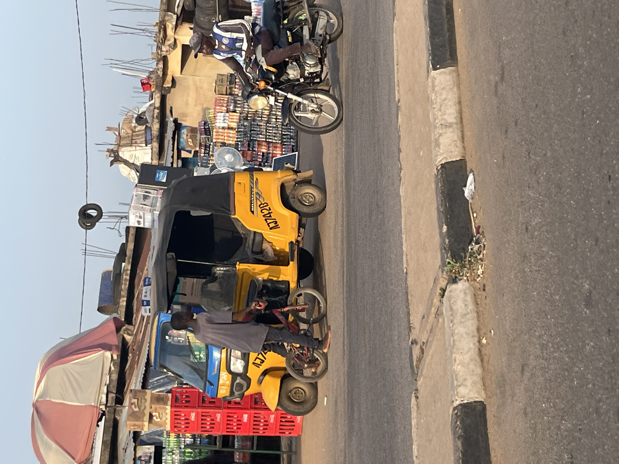 The image depicts a street scene in an urban environment, likely a marketplace or roadside area. In the foreground, a grey asphalt road is visible, bordered by a concrete curb with sections painted black and white. A small patch of weeds grows near the curb.
Dominating the mid-ground are two vehicles. A yellow motorized tricycle, commonly known as an auto-rickshaw, is parked. A young person, dressed in a grey t-shirt and dark pants, is standing next to the tricycle and appears to be interacting with a small red bicycle that is positioned in front of the tricycle. The tricycle has the license plate "AJ7428" visible.
To the right of the tricycle, a man wearing a dark cap, a white and blue vest over a dark shirt, and dark pants is seated on a dark-colored motorcycle. He is looking towards the left of the frame. The motorcycle has a white front wheel and is parked on the road.
In the background, there are various structures that suggest shops or stalls. These include corrugated metal roofs, some of which are rusty. Stacks of red plastic crates are visible to the left. Shelves filled with bottles, likely beverages, are displayed behind the tricycle and on the right side of the image. A large, partially visible umbrella with red and white segments is mounted on one of the structures in the background. The sky above is clear and light blue. The overall impression is a bustling commercial area with a blend of transport and retail activities.