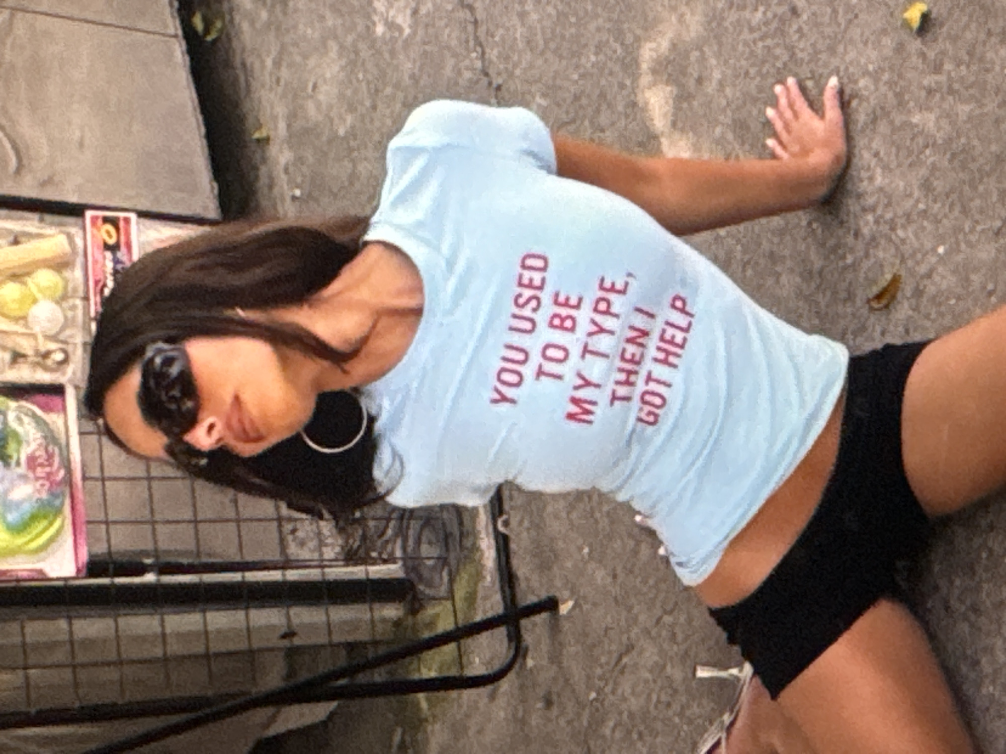 A young woman is lying on her side on a concrete surface, with her back to the camera. She is wearing a light blue t-shirt with red text and black shorts. Her arms are extended, with her right hand resting on the ground. She is wearing large, dark sunglasses and has her hair pulled back. The text on her shirt reads, "YOU USED TO BE MY TYPE, THEN I GOT HELP".
In the background, there is a black metal grate, and to the left of the woman, there are some objects that appear to be on display or for sale. These include a pink box with a colorful image, some yellow spheres, and what looks like a wooden stick. The setting appears to be outdoors, possibly a street or market, with a rough concrete ground and a blurred background of a wall or building. The lighting suggests it is daytime, with natural light illuminating the scene. No specific weather conditions are discernible. The overall impression is a casual, perhaps candid, moment captured in an urban or semi-urban environment. The woman's pose and expression, though partially obscured by sunglasses, do not convey any strong emotions. The location context of Kuta Selatan, Indonesia, suggests a tropical or sub-tropical climate.