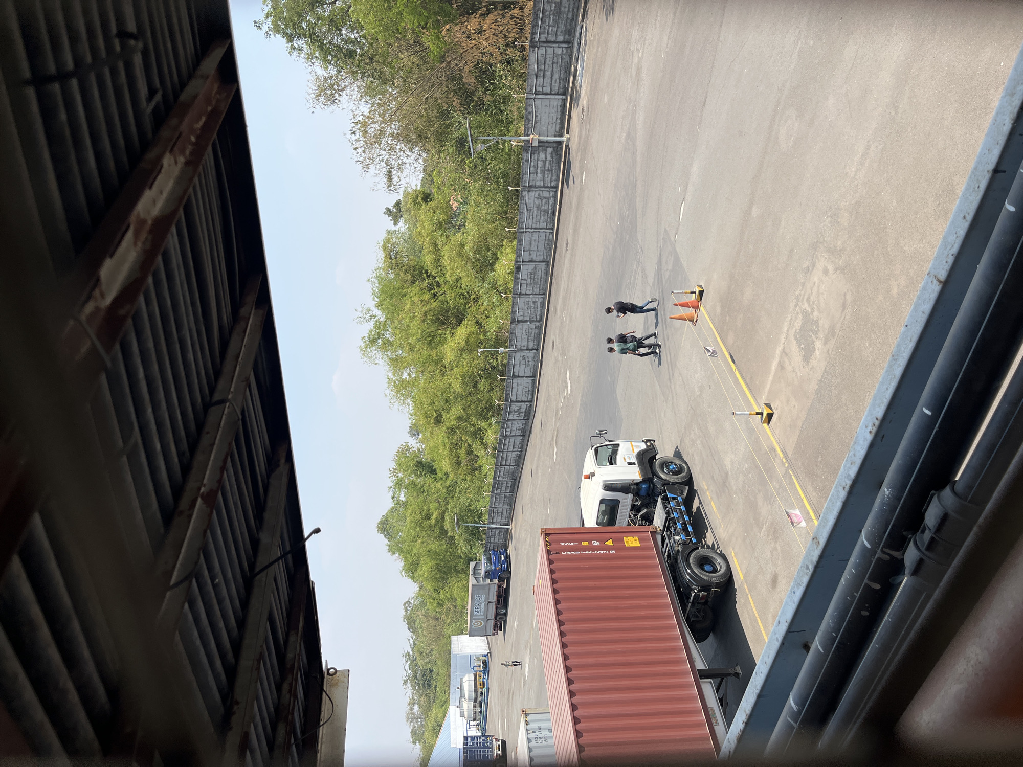 The image shows an outdoor scene at a shipping or logistics yard, possibly located in the city of Pelang, Indonesia. The foreground is dominated by the underside of a structure with a metal roof and exposed beams, framing the view of the yard.
In the middle ground, a large red shipping container is mounted on a white truck with black wheels and blue accents. The truck is parked on an asphalt surface marked with yellow lines. To the left of the truck, a section of another shipping container, appearing to be metallic gray, is visible.
Further into the yard, there is a blue truck with a "SERVICE" logo on its trailer. Behind the trucks, a concrete wall topped with barbed wire runs horizontally across the frame, separating the yard from a dense area of green trees and bamboo. Several lampposts are positioned along the wall.
In the distance, there are white industrial buildings with what appear to be large cooling towers.
Three individuals are walking across the asphalt area in the mid-ground, heading from left to right. Two of them are wearing dark clothing, and one is wearing a green t-shirt. Two orange traffic cones are placed on the asphalt near where they are walking. The sky is clear and pale blue. The overall impression is a functional, operational logistics hub under a bright, sunny sky.