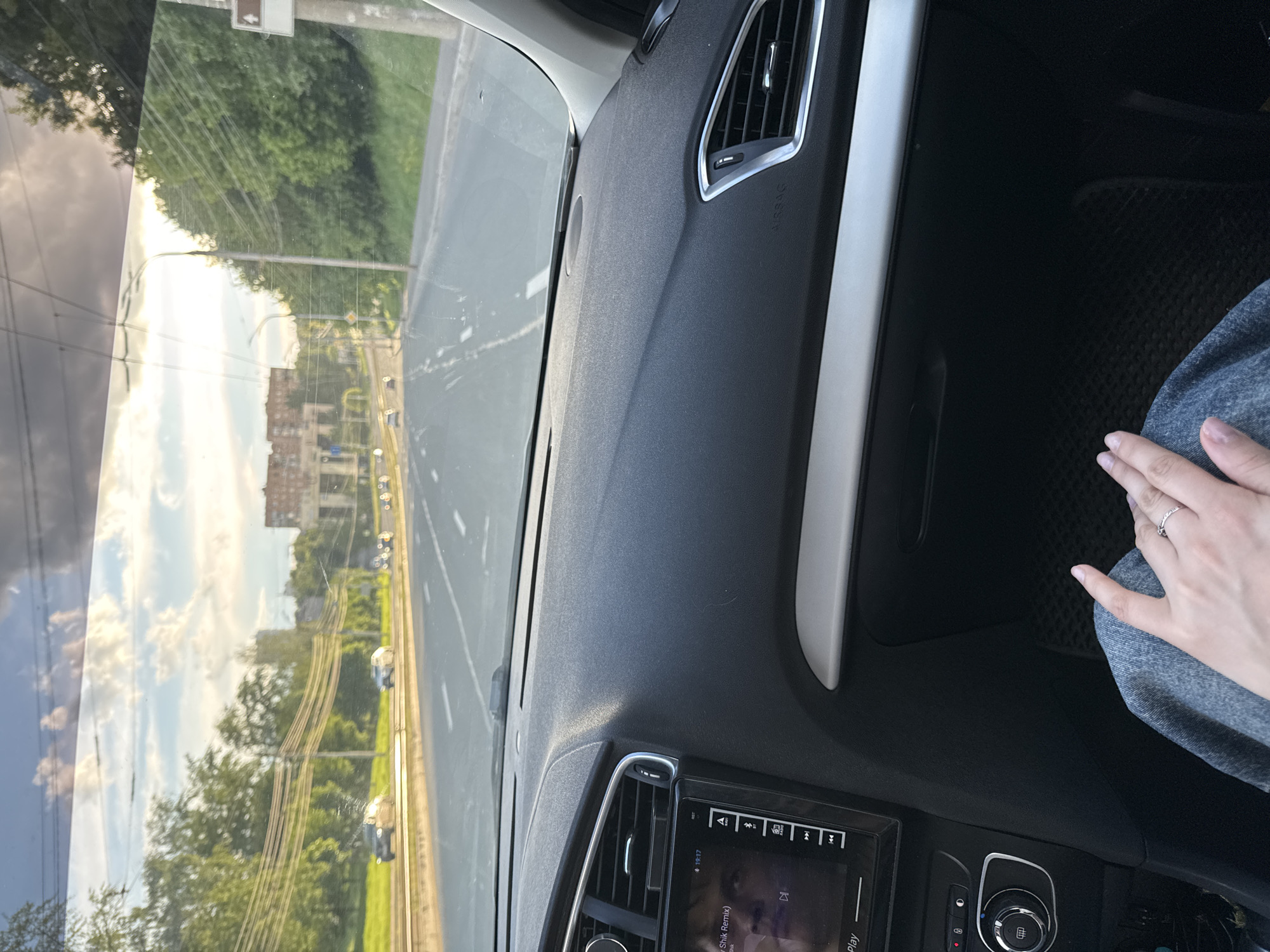 The image shows the interior of a car, specifically the dashboard, with a view of a road and buildings through the windshield. A person's hand rests on their lap, wearing a ring. The road appears to be a multi-lane highway with a grassy median. Buildings are visible in the distance, including what looks like residential or commercial structures. Overhead power lines are present. Based on the architecture and infrastructure visible, as well as the overcast weather, it could be the city of Izhevsk, Russia.