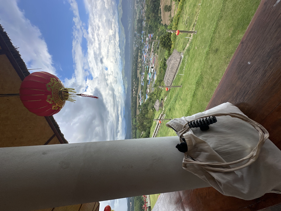 The image displays an elevated vantage point overlooking a valley in Ban Mai Saha Sampan, Thailand. In the immediate foreground, a light-colored cylindrical concrete pillar occupies the left side, beside a dark brown wooden railing or table surface extending horizontally. A light beige fabric tote bag rests on this wooden surface, featuring a black hair claw clip attached to its top edge and a dark object partially visible inside. Suspended from an overhead thatched roof, a prominent red Chinese lantern with gold embellishments and hanging tassels is visible at the top center. A second, partially visible red lantern is located on the far left.
The midground reveals a steep, green grassy hillside descending into the valley. Several evenly spaced wooden posts, each topped with a small red lantern, are positioned along the slope. A small, open-sided gazebo with a thatched roof is visible on the upper part of the hillside, and another building with a thatched roof appears further down the slope. The background showcases a dense cluster of buildings, indicative of a village or small town, situated within the valley, surrounded by agricultural fields and lush vegetation. A chain of mountains and hills defines the distant horizon. The sky above is partially clear blue with significant formations of white and grey cumulus clouds. No individuals are present in the scene.