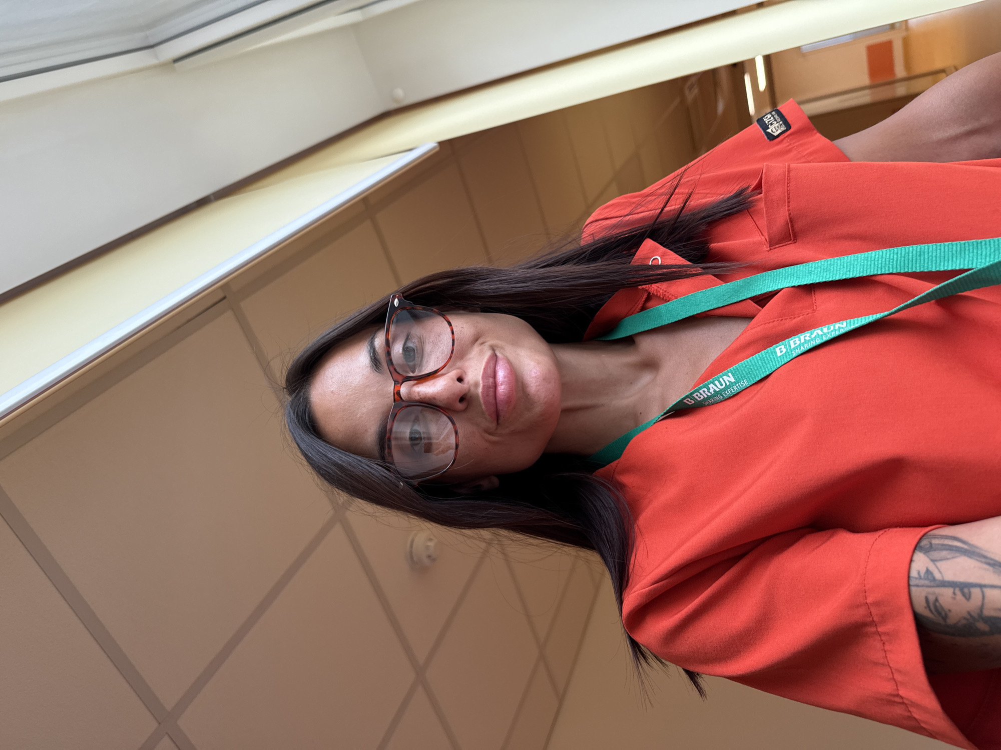 A medium shot captures a woman with dark hair and glasses, wearing a bright orange medical scrub top with a green lanyard around her neck. She has a tattoo on her left arm, partially visible. The background appears to be an indoor hallway with a tiled ceiling and large windows on the right, suggesting a healthcare facility. The image has a shallow depth of field, with the woman in sharp focus and the background slightly blurred. The lighting is natural, coming from the windows. The overall impression is a candid selfie taken in a professional setting. This scene is located in the city of Pleven, Bulgaria.