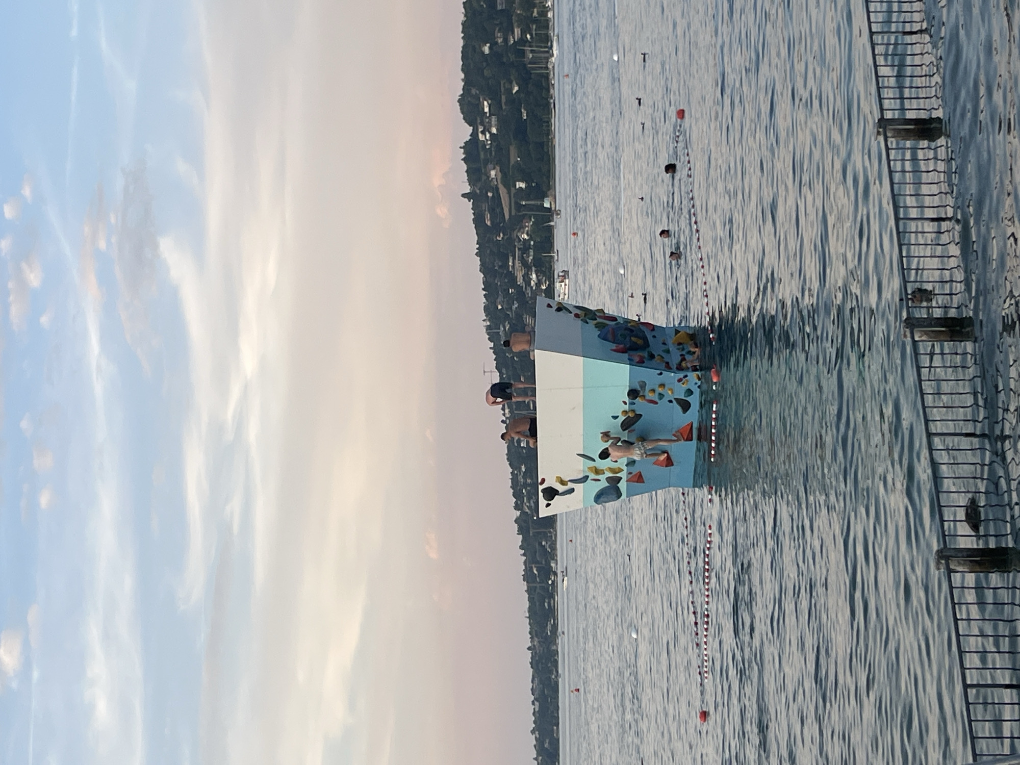 The image depicts an outdoor climbing structure in the water at the Jet d'Eau beach area in Geneva, Switzerland. The structure, resembling a large abstract sculpture painted with colorful geometric shapes, is positioned in the lake. Several individuals are visible on and around the structure. One person is actively climbing the structure. Others are perched on the top, with at least two men sitting or standing. In the surrounding water, several people are swimming, indicated by heads visible above the surface. Buoys and ropes delineate a swimming area. The background shows a tree-lined shore with buildings and boats, indicative of a city environment. The sky is partially cloudy with soft, diffused light, suggesting it is either early morning or late afternoon. A metal fence is in the foreground, with some of its posts submerged in the water.