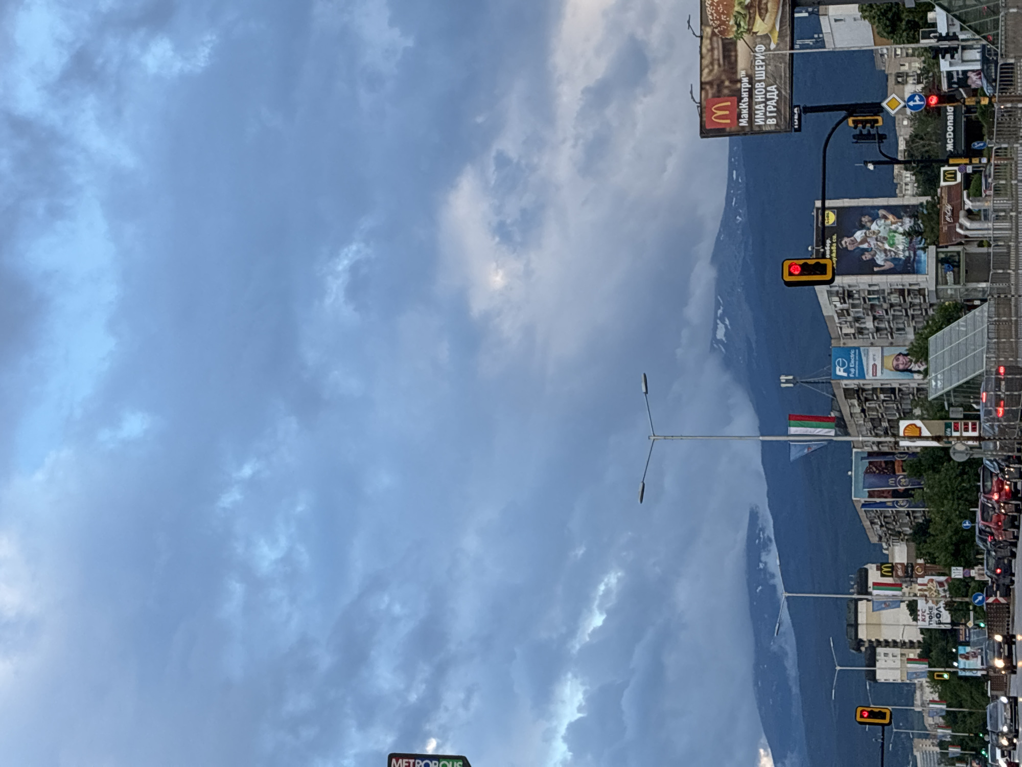 The image, when viewed upright, captures a dynamic urban street scene in Sofia, Bulgaria, set against a majestic mountain backdrop under a cloudy sky. The sky is largely overcast with shades of grey and white clouds, suggesting a cloudy daytime, possibly evening or morning. In the background, a large, dark mountain range, likely Vitosha, dominates the horizon.
The foreground is filled with elements of a busy city street. Numerous commercial buildings line the road, adorned with a variety of billboards and signs. Several street lamp poles and traffic lights are visible; a prominent traffic light in the mid-ground displays a red signal, while another further down shows green.
Multiple brand advertisements and logos are clearly identifiable. A large McDonald's billboard is prominent, featuring a burger and Cyrillic text that translates to "McCountry" and "THERE'S A NEW SHERIFF IN TOWN." Other visible brands include "McDonald's" on a smaller sign, "KFC," "Shell" (logo visible on a building), "Fuji Electric" (with "Fe" logo), and a glimpse of "Lidl" on a sports-themed billboard. Another large sign at the bottom left likely reads "METROPOLIS." A Bulgarian flag (white, green, red horizontal stripes) can be seen flying from a pole, affirming the location context.
The scene conveys the sense of a bustling urban environment with ongoing traffic and commercial activity, contrasted with the natural grandeur of the mountains and the changing weather overhead.