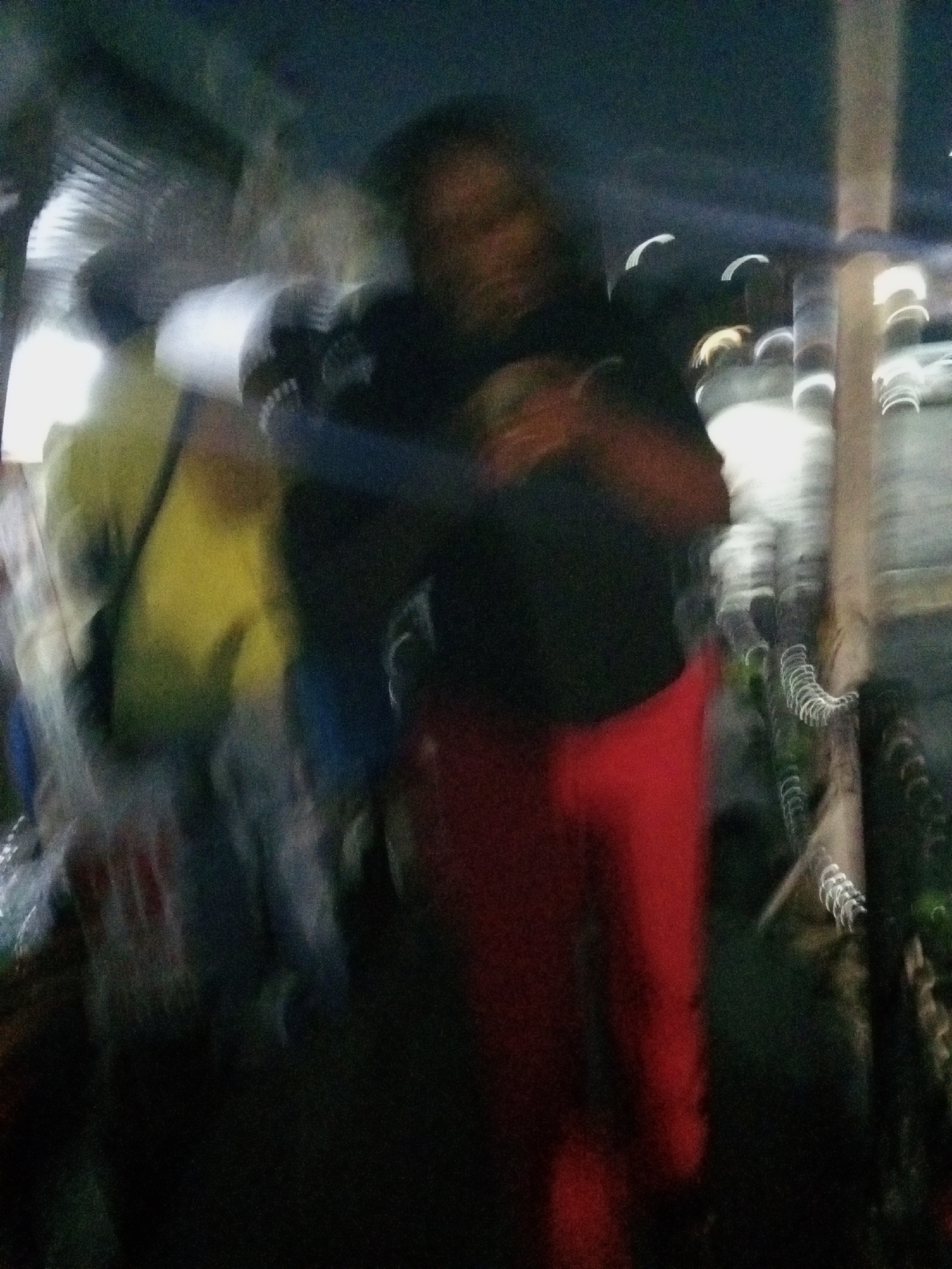 The image depicts a blurry scene at night in Ado, Nigeria. A person wearing red pants and a dark top is the main subject. Another person wearing a yellow shirt is visible to the left, along with what appears to be merchandise or goods, indicating a possible market or street vending environment. Light sources create curved streaks, likely from vehicles or other city lights, further suggesting a nighttime setting. A vertical, possibly wooden, structure is also present on the right.
