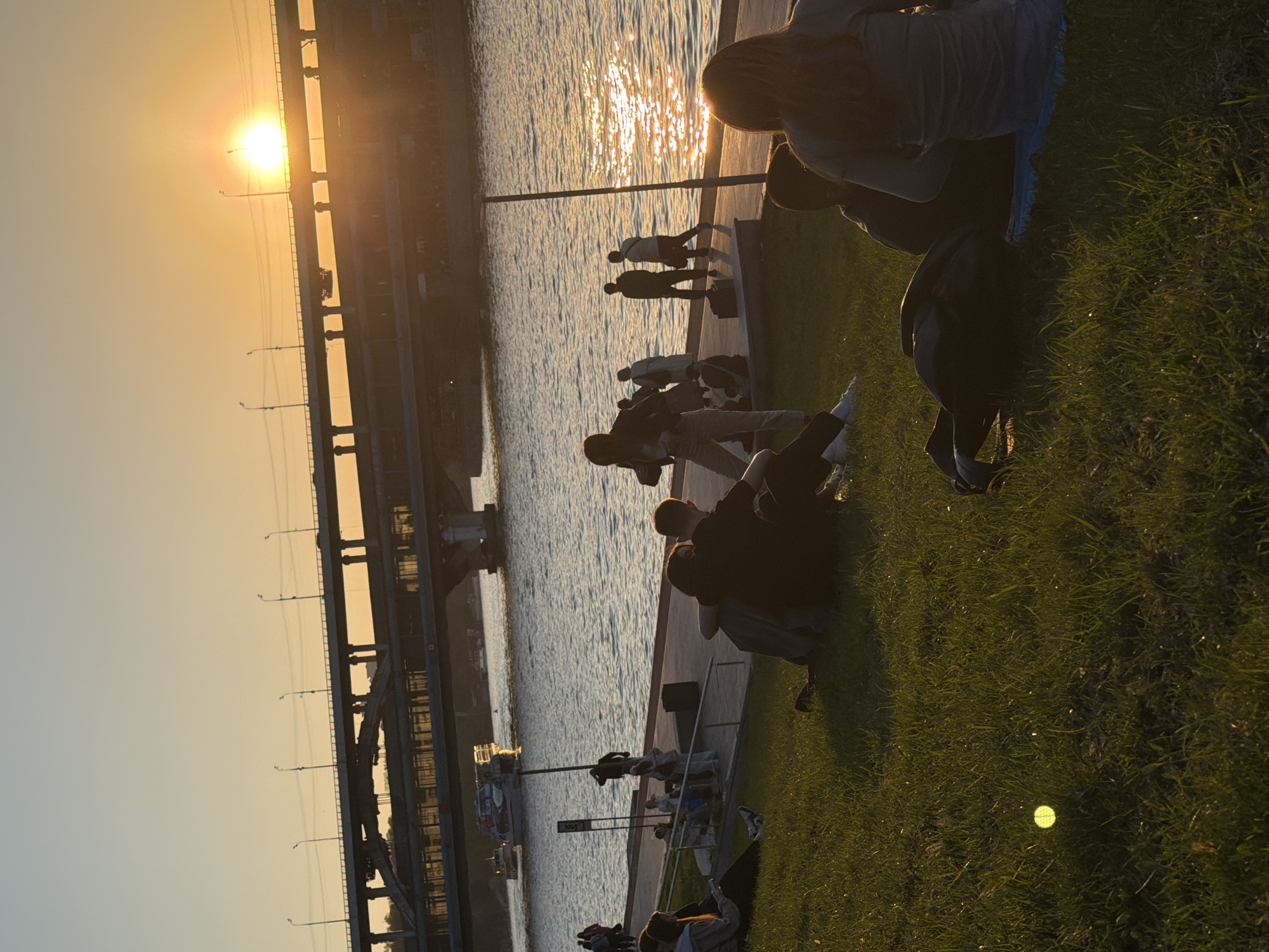 The image depicts a cityscape at sunset, likely in Moscow, Russia, given the architectural style of the bridge on the left. The sun is low on the horizon, creating a golden light and reflecting off the surface of the river. A bridge structure spans the river, its metal girders visible against the bright sky. Along the riverbank, people are gathered on a grassy area. Some are sitting and relaxing, while others are walking along the edge of the water. The focus is on the human element interacting with the urban landscape at the end of the day.