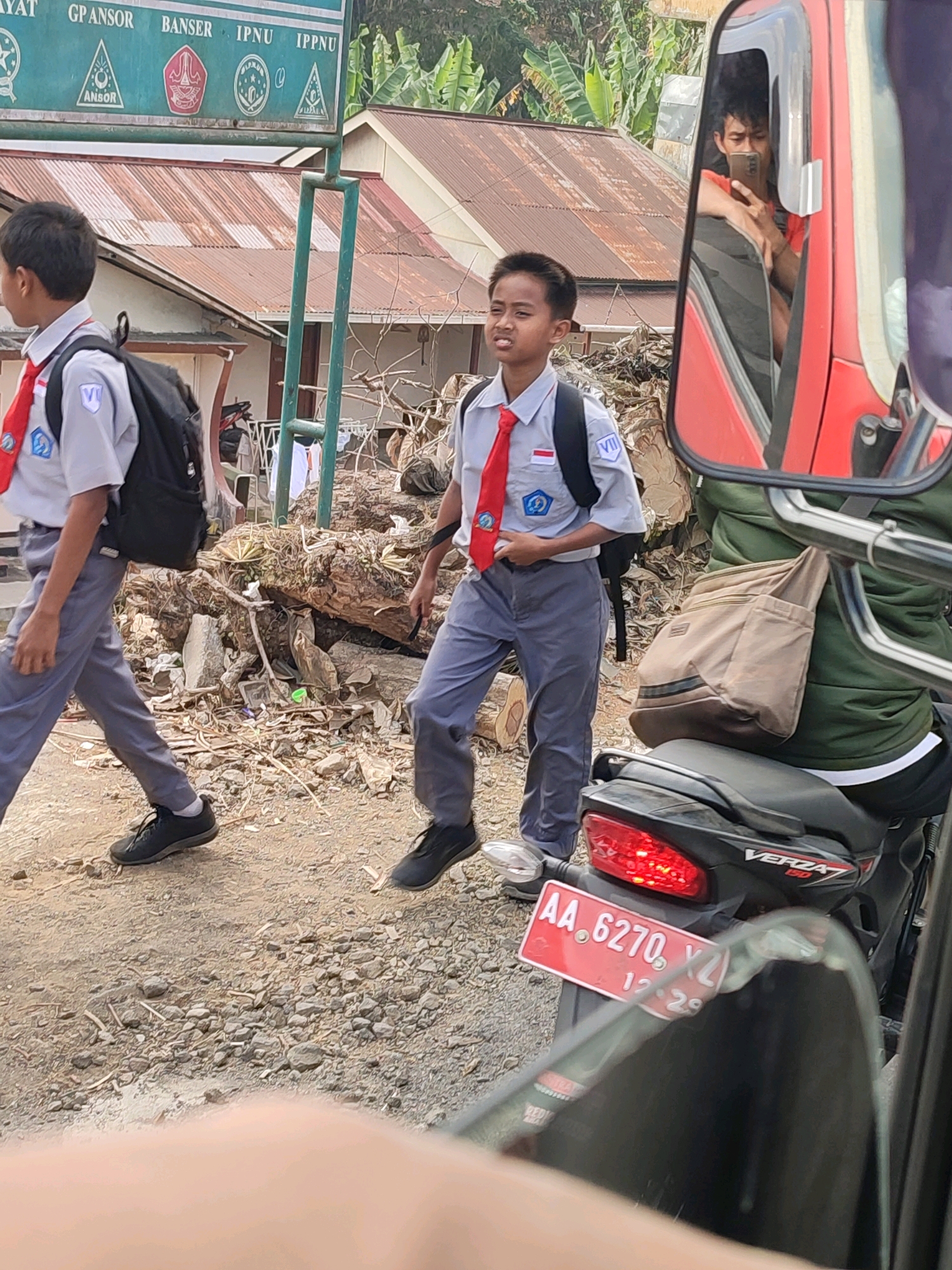 Two young Indonesian boys in school uniforms, gray shirts with red ties and gray pants, walk on a dirt road in front of a building with a corrugated metal roof. The boy on the left carries a black backpack. The boy on the right looks towards the camera with a grimace. A red side mirror of a vehicle reflects a young man holding a smartphone. A black motorcycle with a red license plate is parked on the right side of the image. The license plate reads "AA. 6270. YZ". The background shows trees and a sign with Indonesian text. The environment appears to be rural.