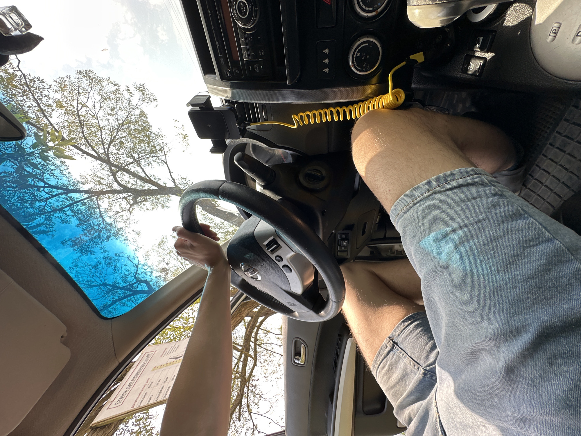 A first-person perspective shot shows a person's legs and hands inside a vehicle. The individual is wearing denim shorts and is holding the steering wheel. The car's interior is visible, including the dashboard, a phone mount with a coiled yellow charging cable, and a portion of the car's center console. Through the windshield, trees and a bright sky are visible. A sign with text is also attached to the windshield. The image was taken in Makiv, Ukraine.