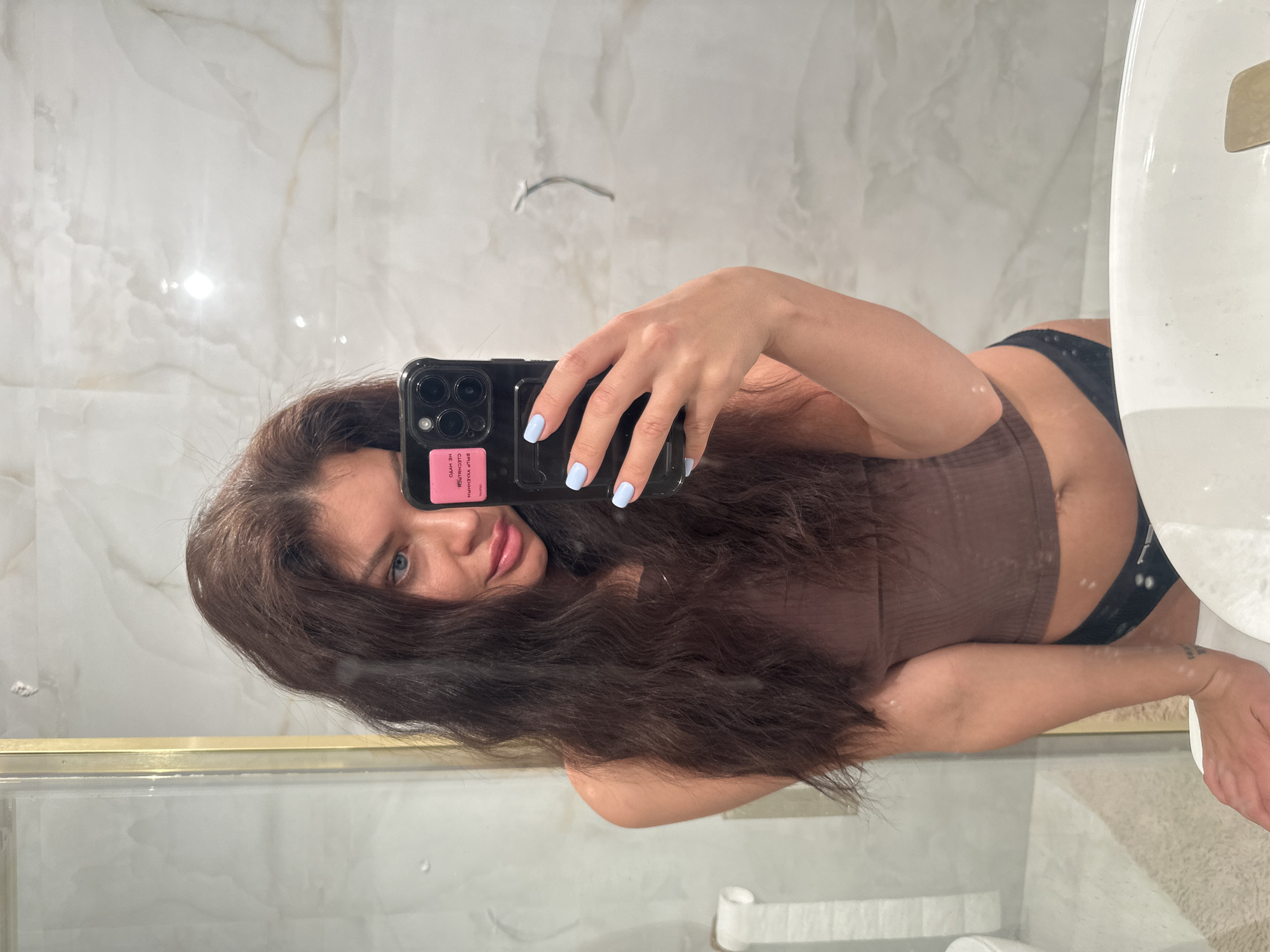 A woman with long, dark brown hair and blue eyes is taking a selfie in a bathroom mirror. She is wearing a brown ribbed tank top and black underwear. The bathroom has white marble walls and a toilet is visible in the background. The location cannot be determined from the image alone. There is no definitive evidence of Khimki, Russia.