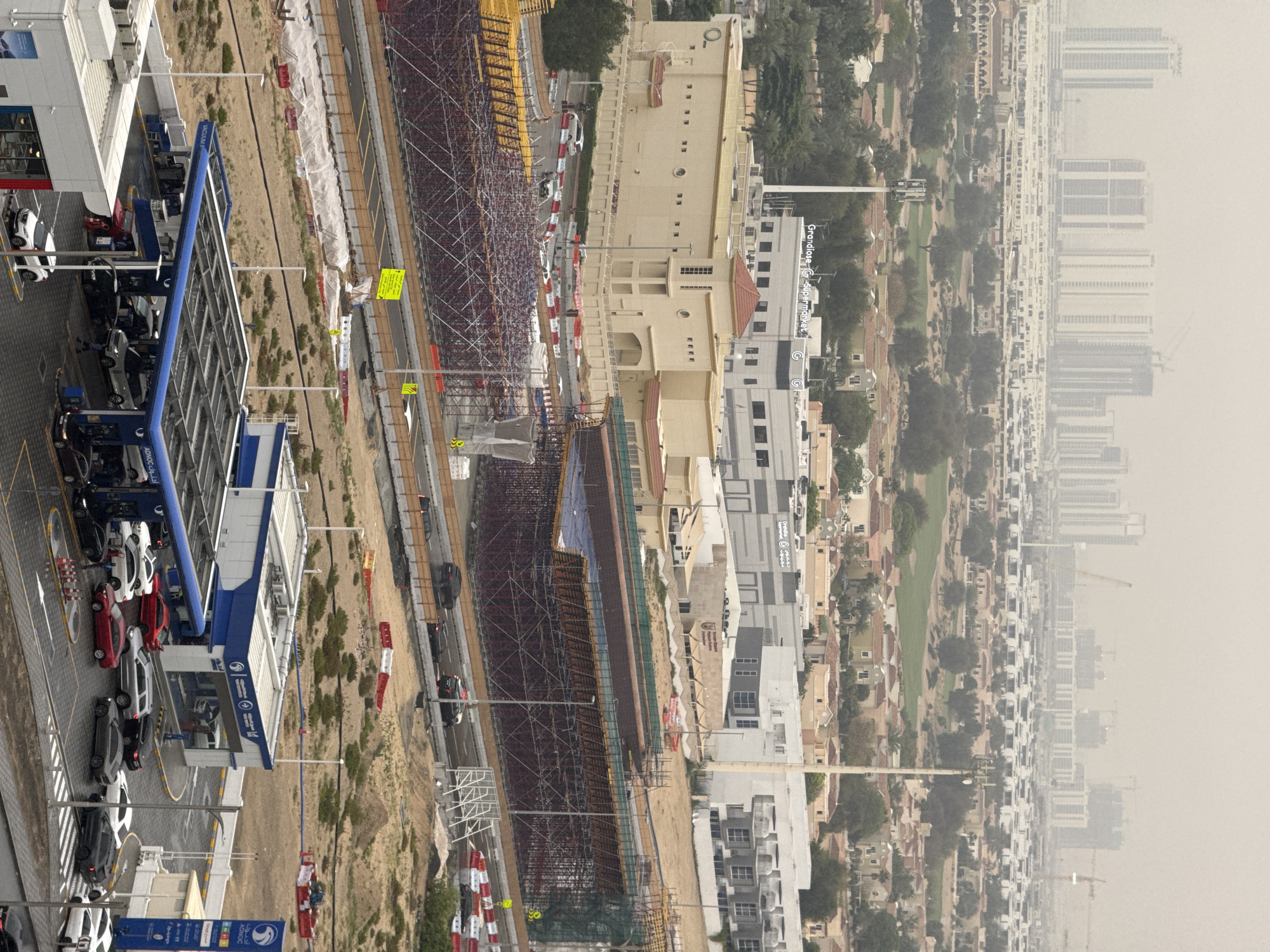 The image depicts an aerial view of a section of Dubai, United Arab Emirates. An ADNOC service station is in the foreground, with several cars parked at the pumps. Beyond the service station, there is a construction site with extensive scaffolding and ongoing building activity. Adjacent to the construction site are residential buildings, followed by a golf course. In the distance, a hazy skyline shows more high-rise buildings. The overall impression is a mix of urban development, infrastructure, and residential areas within the city.