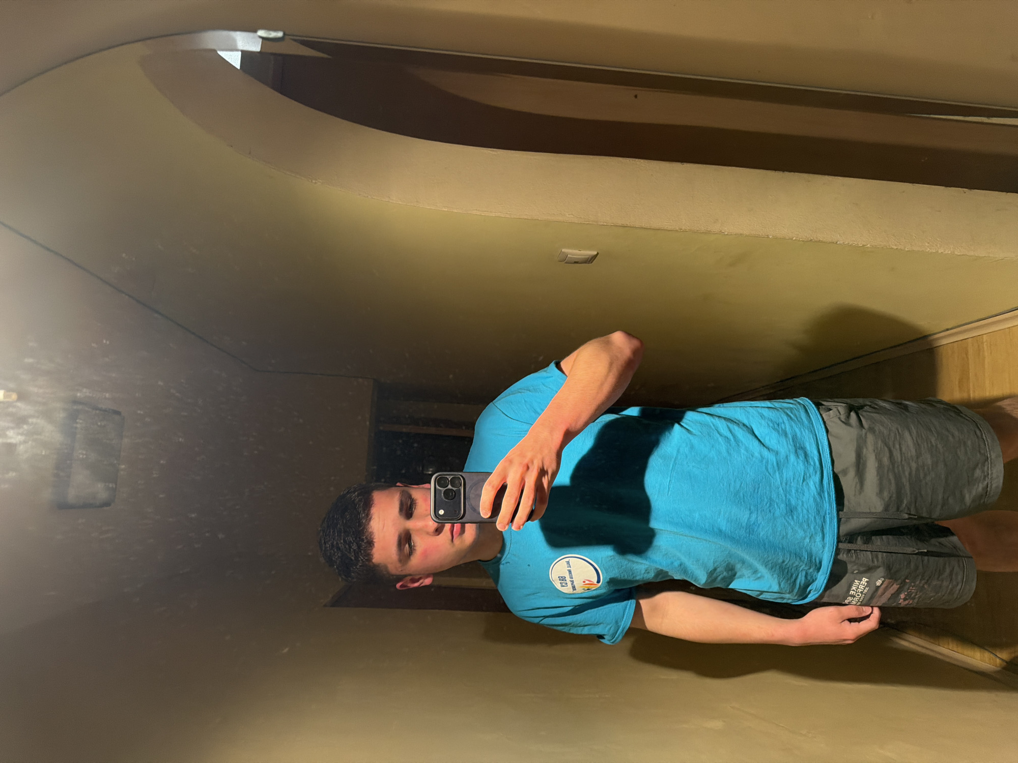 A young man is taking a selfie in a mirror. He is wearing a blue t-shirt and gray shorts. The environment appears to be an interior hallway or room with a curved ceiling. The location is not identifiable as Sofia, Bulgaria.