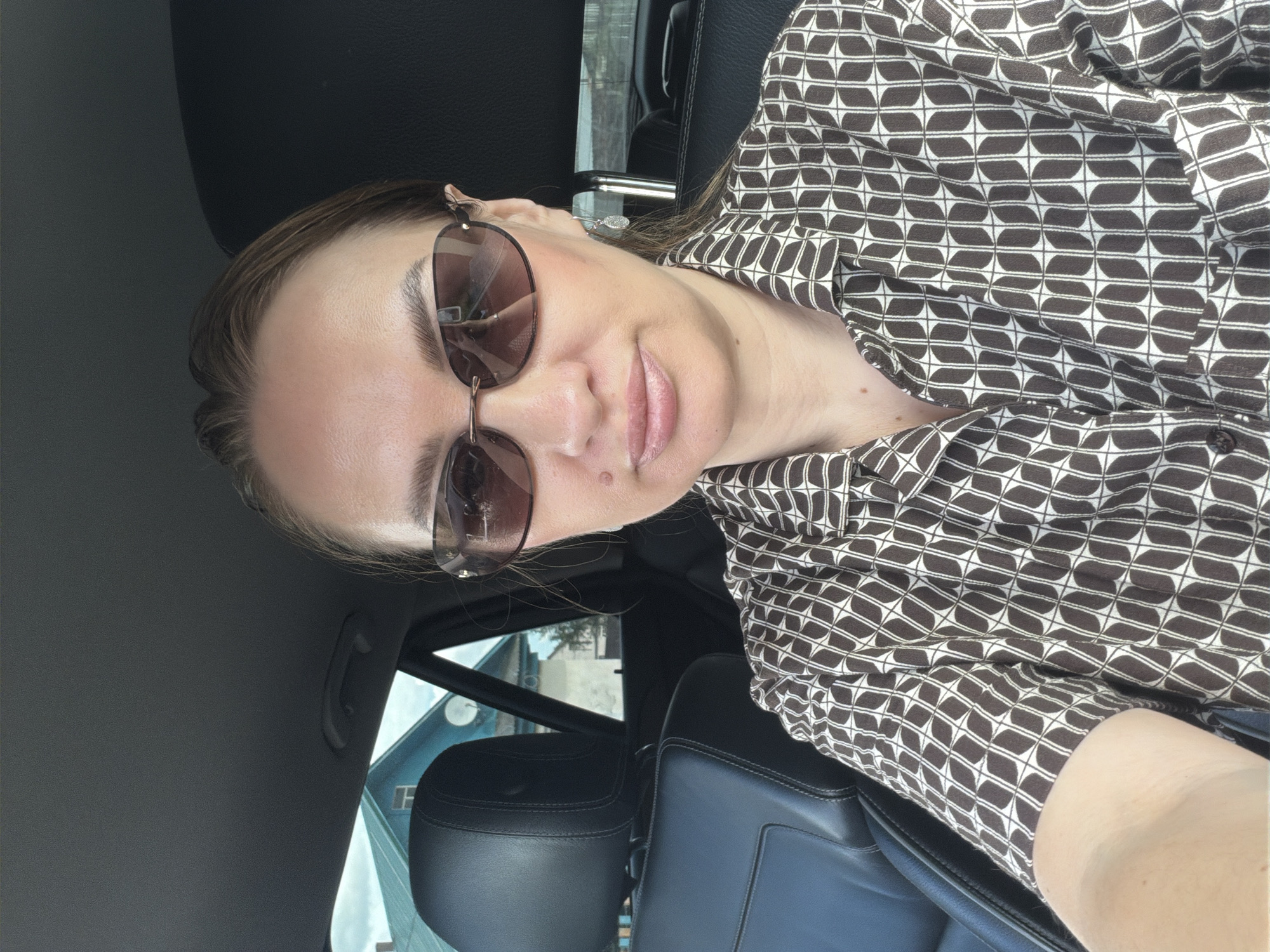 A woman wearing sunglasses and a brown and white patterned shirt is seated in the driver's seat of a car. She is looking towards the camera. The interior of the car is visible, with black leather seats. Outside the car window, a house with a blue roof and a satellite dish is visible. The scene is set in Mezhevoy, Russia.