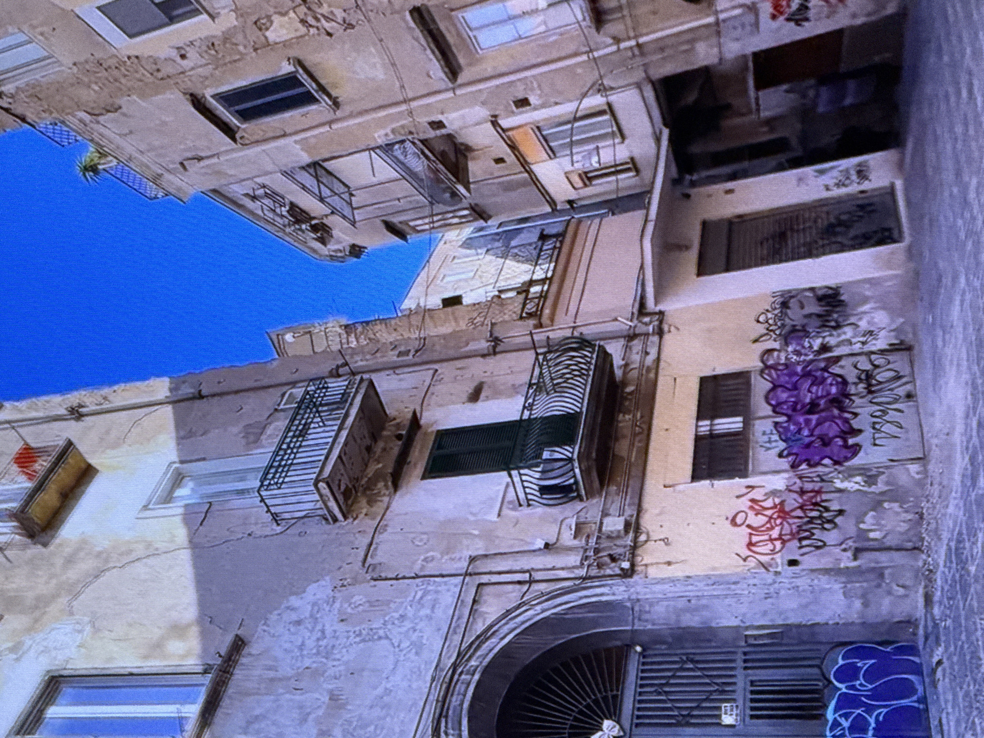 This media file depicts an outdoor scene looking up at the sides of multiple aged, multi-story buildings, likely an apartment complex. The buildings are constructed from weathered concrete with visible cracks and peeling paint, showing signs of disrepair. Many windows are present, some with metal bars for security, and some with shutters, including a green shuttered window. Several balconies with decorative iron railings jut out from the structures. The architecture suggests an older urban setting.
The buildings are adorned with graffiti in various colors, including purple, red, and blue, covering portions of the walls and some windows. The sky visible in the upper left quadrant is a bright, clear blue, indicating it is daytime and the weather is likely sunny and fair. Electrical conduits and wiring are visible running along the exterior walls of the buildings. The image appears to be taken from a low angle, looking upwards, giving a sense of the height and density of the buildings.
There are no people clearly visible in the image. The overall impression is one of urban decay and the presence of street art in a densely populated area. The location context provided is Dubai, United Arab Emirates, which might seem unusual for the style of architecture and apparent wear, but the description is based solely on the visual elements present. No discernible text is legible within the graffiti.