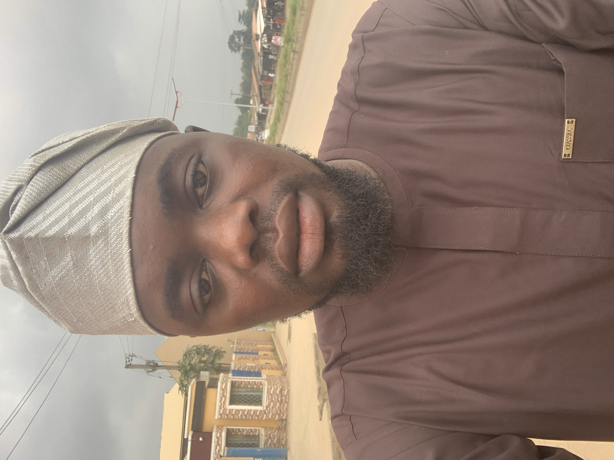 A close-up, front-facing selfie shows a man in Shagamu, Nigeria, with a grey, patterned cap. He wears a brown, long-sleeved traditional outfit with a subtle seam down the front. A small, gold-colored tag with the word "CIASSLC" is attached to the left chest pocket. He has a beard and is looking slightly to his right. In the background, a street is visible with buildings, power lines, and trees. The sky is overcast. The image appears to have been taken outdoors, possibly on a street or road in a residential or commercial area.