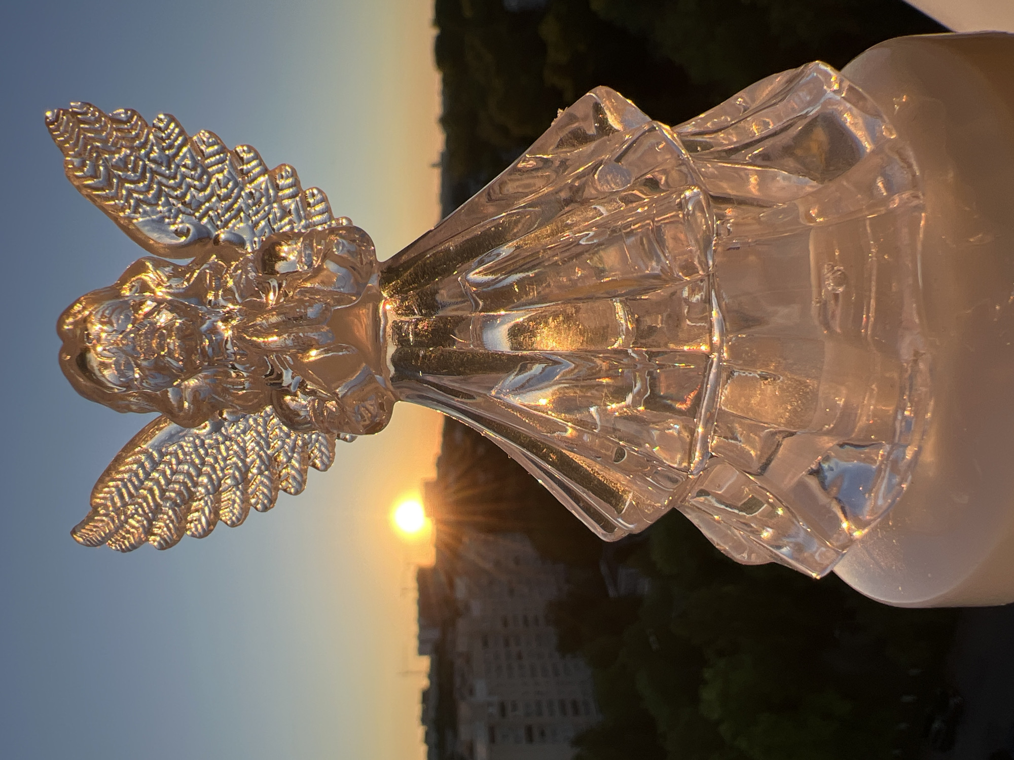 An angel figurine made of clear, faceted glass is positioned in the foreground, illuminated by the setting sun. The figurine has delicate wings and a flowing gown, catching the light and creating prismatic effects. Behind the figurine, the sun is low on the horizon, casting a warm glow over the scene. Silhouetted buildings and trees suggest an urban environment, likely in Minsk, Belarus, given the context of the request. The sky is a gradient of orange and blue, transitioning from the warm hues of sunset to the cooler tones of twilight. The overall impression is one of serene beauty and tranquility, with the glass angel acting as a focal point against the backdrop of the urban sunset.