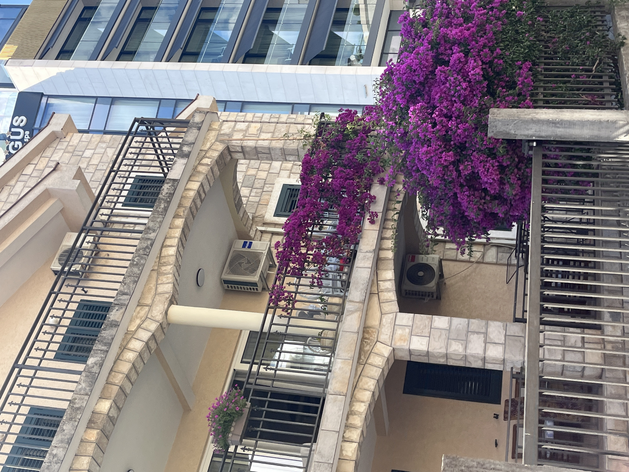 The image depicts the exterior of a building with a vibrant display of bougainvillea flowers in Budva, Montenegro. The building features balconies with black metal railings and stone accents. An air conditioning unit is visible on one of the balconies. Adjacent to this building is a modern glass-facade building with multiple levels and balconies. The foreground shows a metal fence. The scene suggests a residential or hotel complex in a sunny location.