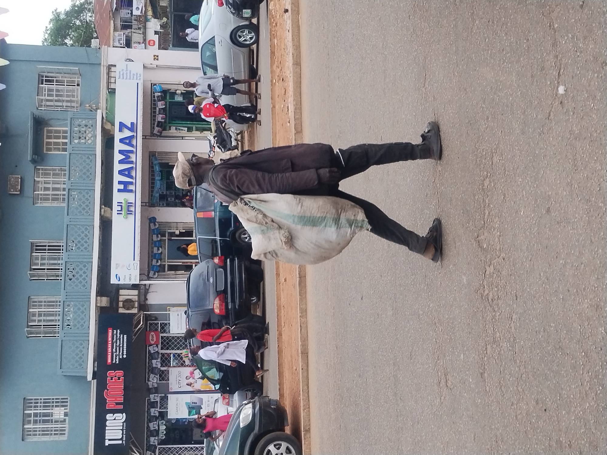 A man carries a large sack on his back while walking on a paved road in Jos, Nigeria. The man is wearing dark clothing and a light-colored hat. Several cars are parked along the side of the road and people are walking along the sidewalk. The businesses "Twins Phones" and "Hamaz" are visible.