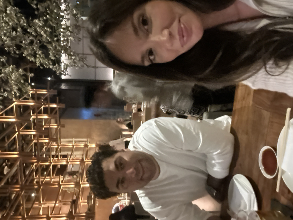 A man and a woman pose for a selfie at a restaurant. The woman has long, dark hair and a striped shirt. The man, in a white t-shirt, smiles at the camera. A wooden table is partially visible with a small bowl of sauce, a plate, and chopsticks. Behind them, there's a decorative structure with a grid-like pattern, and trees are visible outside. There are other people visible in the background, appearing out of focus.
