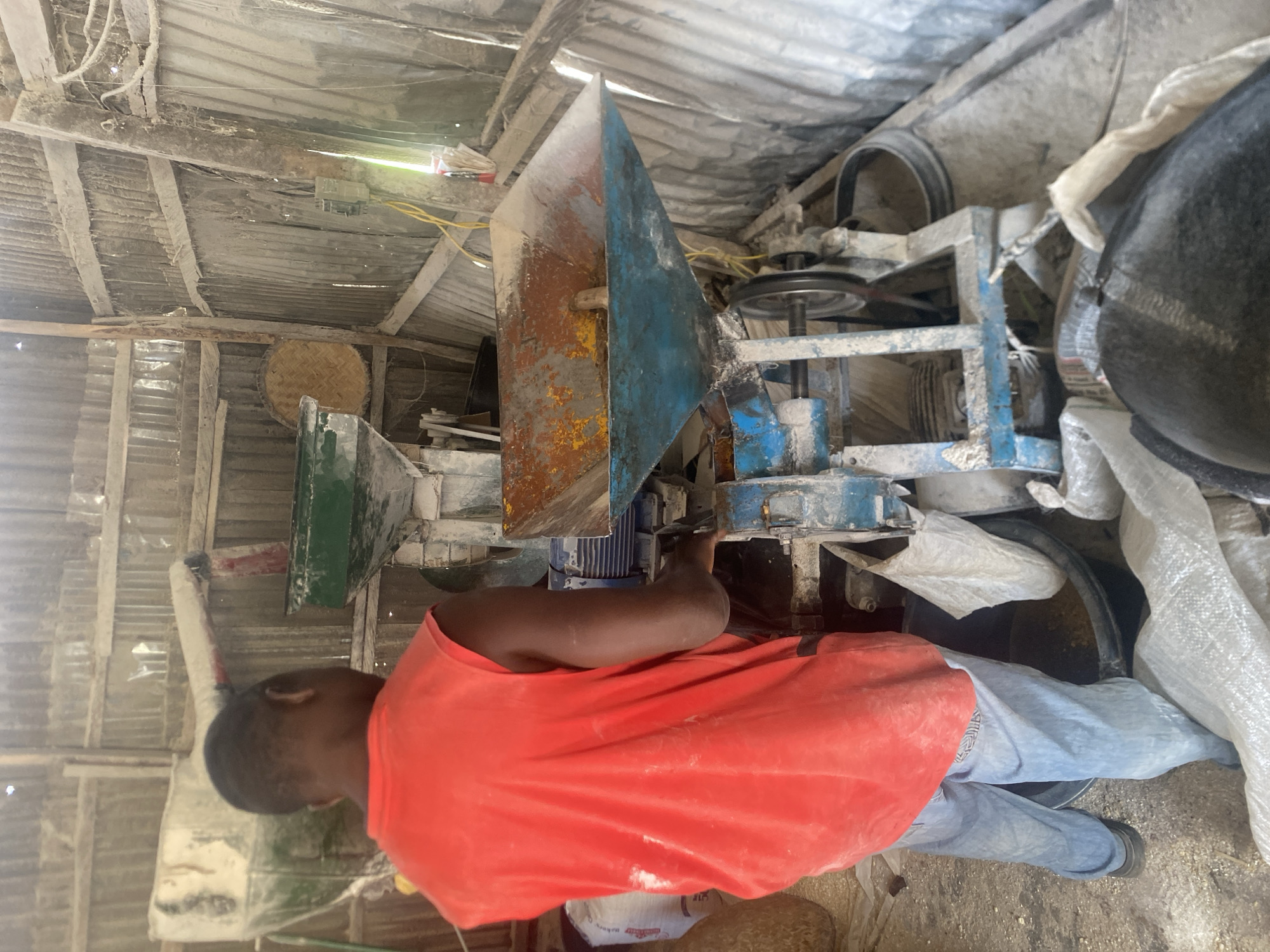 In Bukuru, Nigeria, a man in a red sleeveless shirt and blue jeans operates a milling machine. The machine, likely used for processing grains, is a dusty blue metal structure with hoppers and a rotating belt system. A portion of a blue motor is visible. The man's hands are near the machine's controls. Surrounding the equipment are sacks of grain or feed, some open and some sealed, and large black buckets. The environment appears to be an open-air structure with corrugated metal walls and wooden supports, covered in dust. A woven basket is visible on the wall in the background.