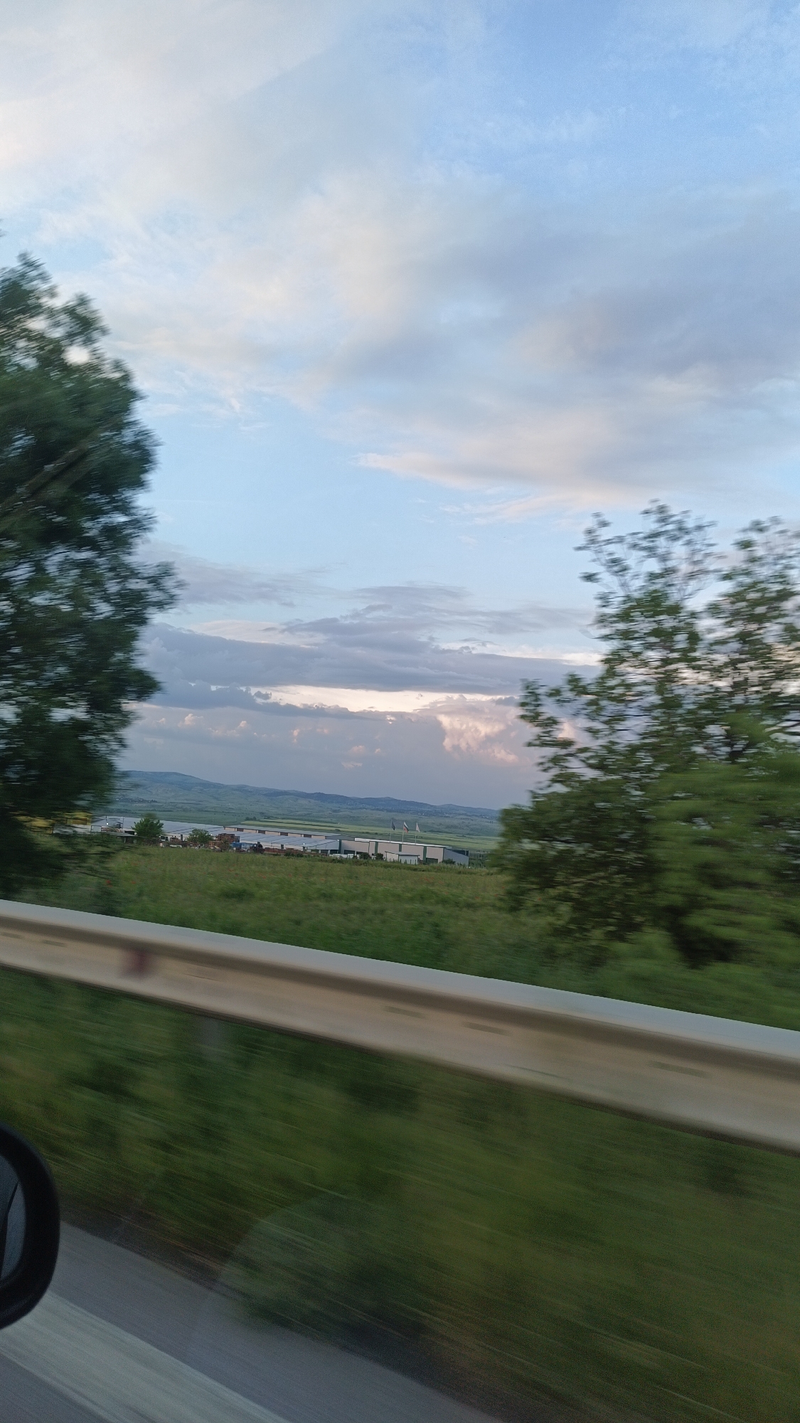 The image depicts a view from a moving vehicle, possibly a car. The foreground is blurred due to motion. Visible through the windshield are trees on both sides of the frame. A metal guardrail is in the lower portion of the image. Beyond the trees and guardrail, a field or grassy area extends into the midground. In the distance are low rolling hills or mountains under a cloudy sky. Several low, white, industrial-type buildings with flat roofs are visible in the midground. Two flags are waving in the background.
There is no information within the image that definitively places it in the city of Burgas, Bulgaria. Determining the location based solely on the provided image would require further information, such as landmarks, unique architectural details, or signage specific to that area.