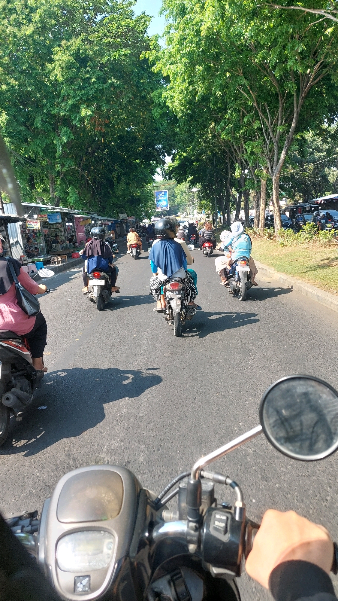 The image depicts a street scene in Sepande, Indonesia, likely during the daytime given the bright sunlight and shadows. The road is lined with lush green trees on both sides, creating a shaded canopy. On the left side of the road, there are several small stalls or shops with colorful awnings. The road itself is paved and appears to be a busy thoroughfare, with multiple motorcycles navigating the traffic.
In the foreground, the perspective is from the rider of a motorcycle, showing the handlebars, a rearview mirror, and the rider's hand. Several other motorcycles are visible in front, most carrying two people, including women wearing headscarves and helmets. One motorcycle in the mid-ground has a license plate visible, reading "26B". The people on the motorcycles are generally wearing casual clothing. The overall atmosphere suggests a typical day of commuting or travel in a bustling Indonesian town.
