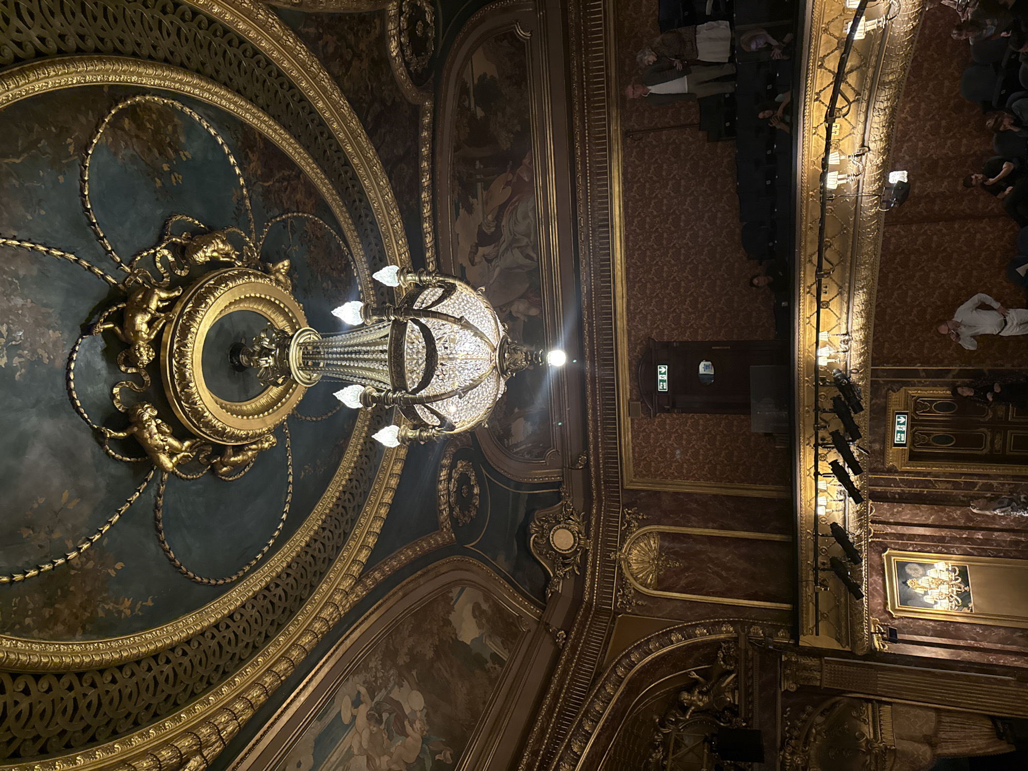 The image captures the opulent interior of a theatre, likely the Royal Opera House in London, United Kingdom, viewed from a vantage point looking up. The ceiling is adorned with intricate gold detailing, including concentric circles, decorative carvings, and painted murals. A grand chandelier hangs suspended from the ceiling, illuminating the space. The theatre's boxes are visible along the right side of the frame, with a few audience members seated within them. The overall atmosphere is one of classical elegance and grandeur.
