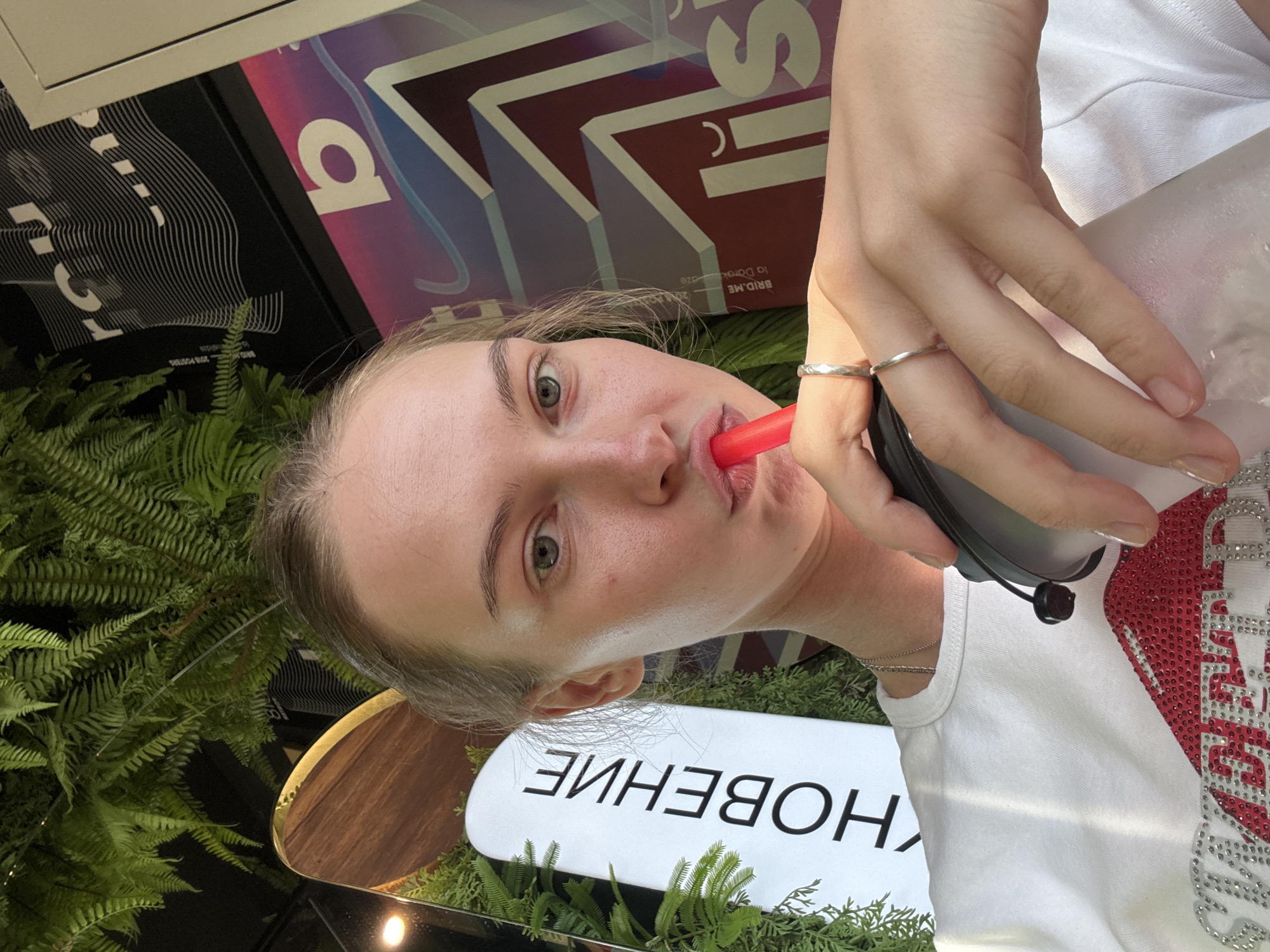 A young woman with blonde hair pulled back is taking a selfie while holding a drink with a red straw. She is wearing a white t-shirt adorned with red and silver rhinestones. Her left hand, wearing two silver rings, holds the cup. In the background, there are lush green ferns and a sign with Cyrillic text that reads "ОБНОВЛЕНИЕ". Behind that, there are graphic posters with abstract designs and lettering. The overall ambiance suggests an indoor setting with vibrant, modern decor, possibly a cafe or a trendy establishment in the city of Perm, Russia.