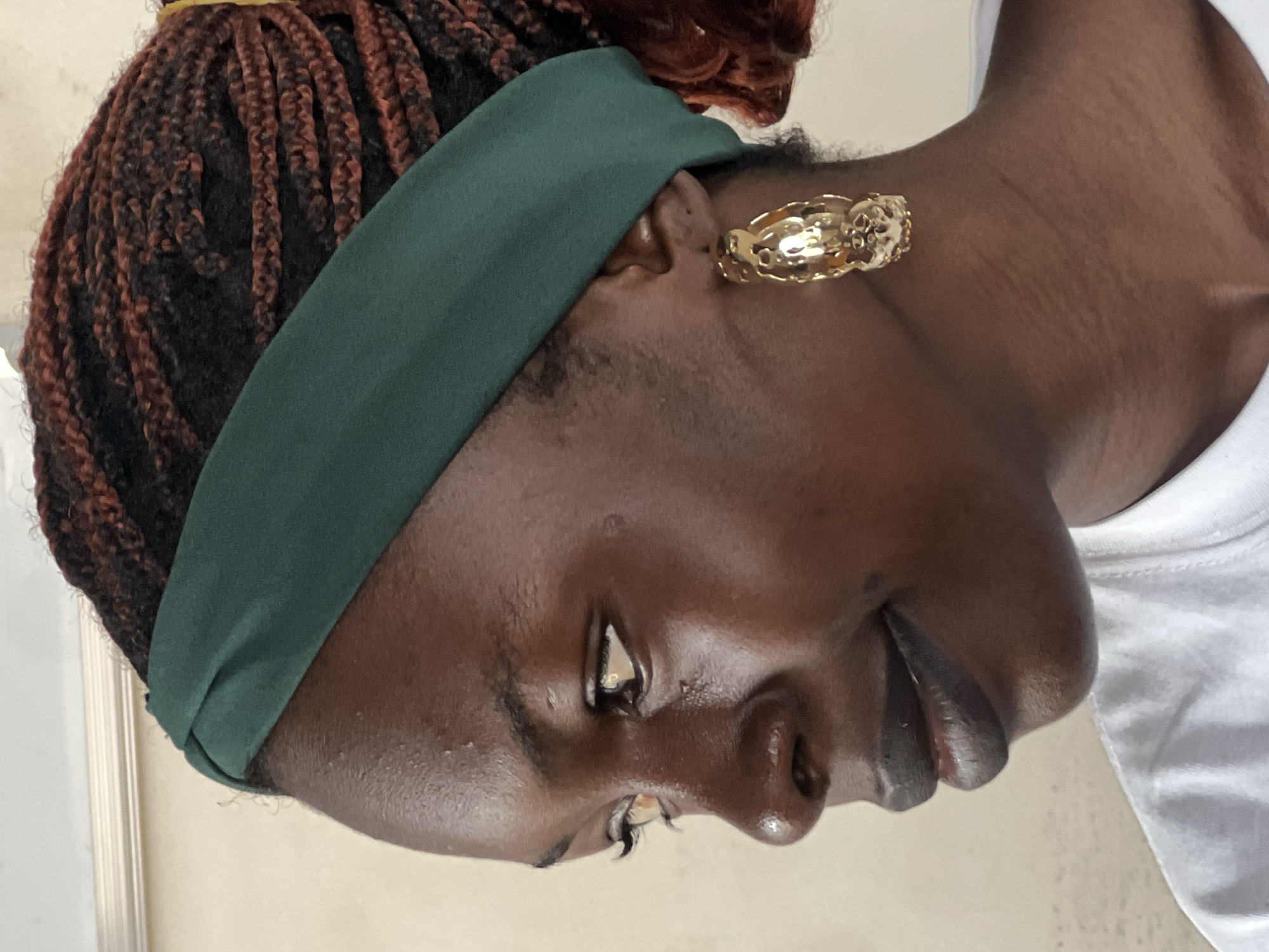 A close-up, profile view shows a young Black woman wearing a dark green headband and large gold hoop earrings. Her hair is styled in braids with reddish-brown highlights. She wears dark lipstick and a white shirt. The background is a plain, light-colored wall. The image is sharp and well-lit, with the woman's skin appearing dewy. No specific details about the city of Jos, Nigeria, are visible in the image.