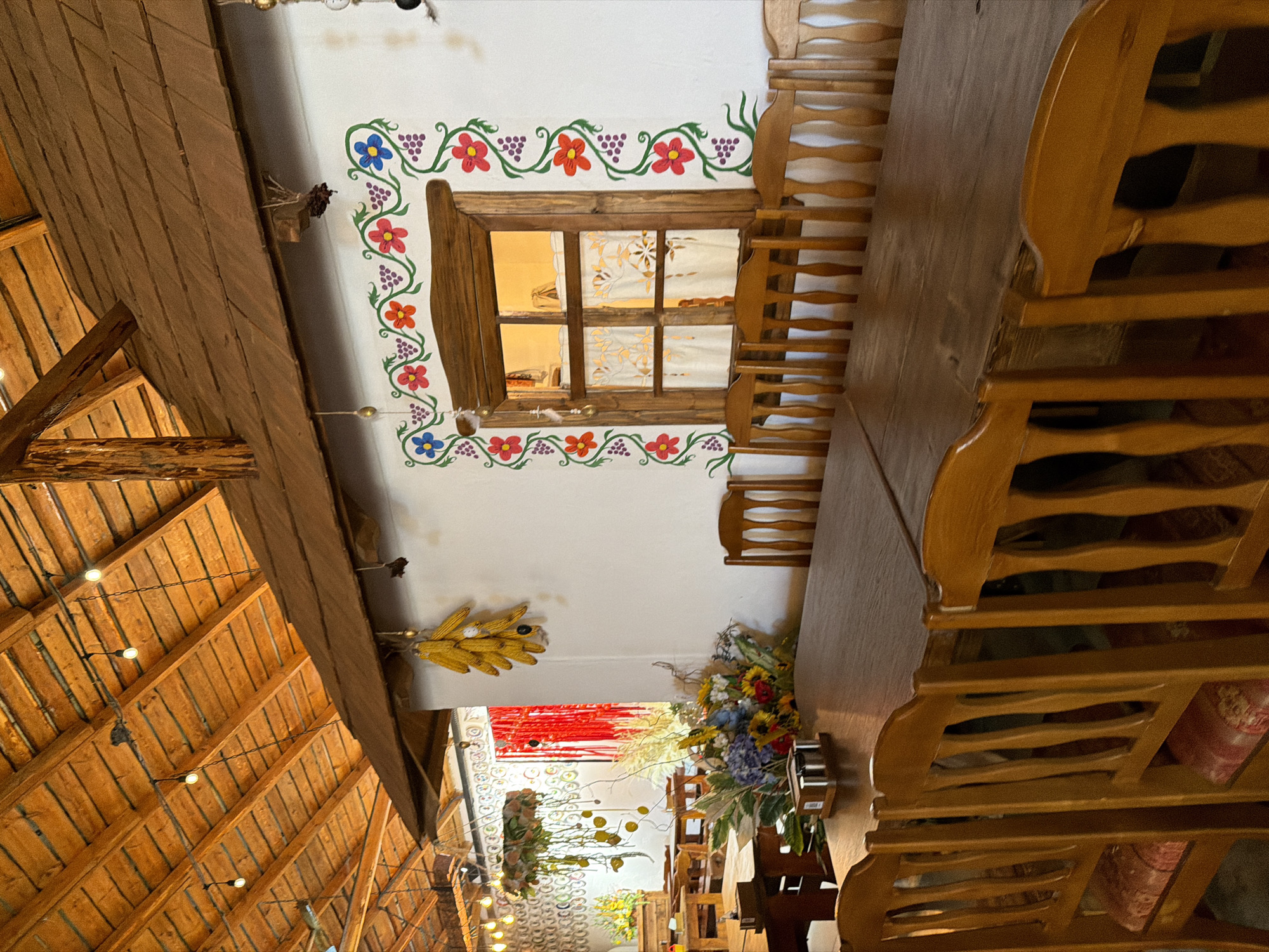 The image depicts the interior of a rustic dining area, likely a restaurant or guesthouse, with a traditional Ukrainian theme. A long, wooden table with several wooden chairs surrounds it occupies the foreground. The table is set with a salt and pepper shaker set. In the background, a window with a wooden frame is adorned with colorful floral and vine-like painted decorations. Above the window, dried corn cobs are hanging as a decorative element. The ceiling features exposed wooden beams and simple light fixtures. The overall ambiance suggests a cozy and authentic setting in Velykyi Khodachkiv, Ukraine, possibly a rural establishment.