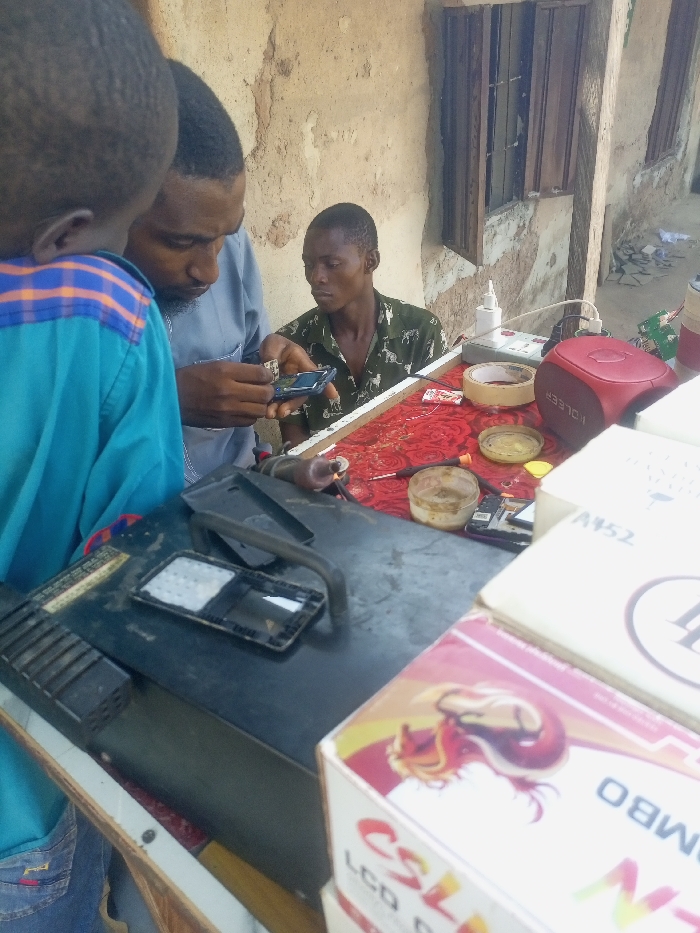 The image depicts an outdoor or semi-outdoor mobile phone repair scene in Akwanga, Nigeria. Three male individuals are present. In the center, an adult male with a beard, wearing a light-colored long-sleeved shirt, actively manipulates the internal components of a disassembled smartphone held in his left hand. To his immediate right, a second adult male with short hair, wearing a green camouflage-patterned shirt, is seated and attentively observes the repair process. Partially visible in the extreme foreground on the left is the back of a third individual, likely a child or young person, wearing a blue shirt with orange and white shoulder stripes, also observing.
The repair is conducted on a two-part workstation. The immediate foreground surface is dark and holds a disassembled phone frame or screen, along with a black curved tool and a rectangular device. The background work surface is covered with a red patterned cloth, displaying various repair items: a red HOLEER-branded device, a roll of masking tape, small open containers with yellowish residue, electronic components, a white bottle, and a small screwdriver. A stack of white cardboard boxes, some labeled "CSL LCD," is visible in the extreme right foreground. The background consists of a weathered, light-colored wall with two barred windows.